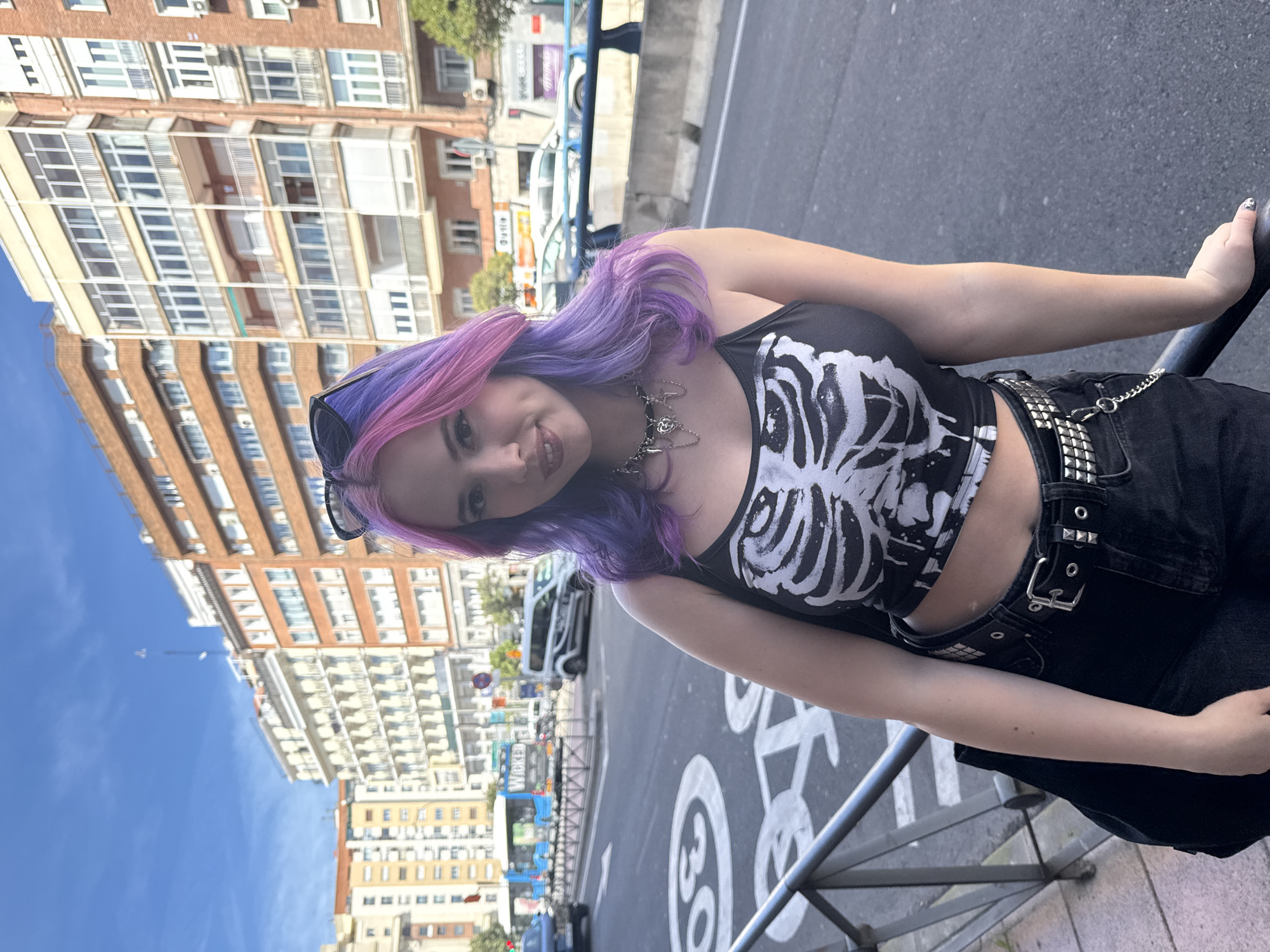 A young woman with dyed purple and pink hair stands on a street in Madrid, Spain. She wears a black tank top with a white skeleton design, black pants with a studded belt, and sunglasses resting on her head. She leans against a metal railing, smiling at the camera. Behind her, a street runs parallel to the railing, with buildings of varied heights and architectural styles lining the other side. A bus and other vehicles are visible on the street. Street signs, including a traffic sign marked with the number 30, are also present. The sky is blue, with a few wispy clouds visible.