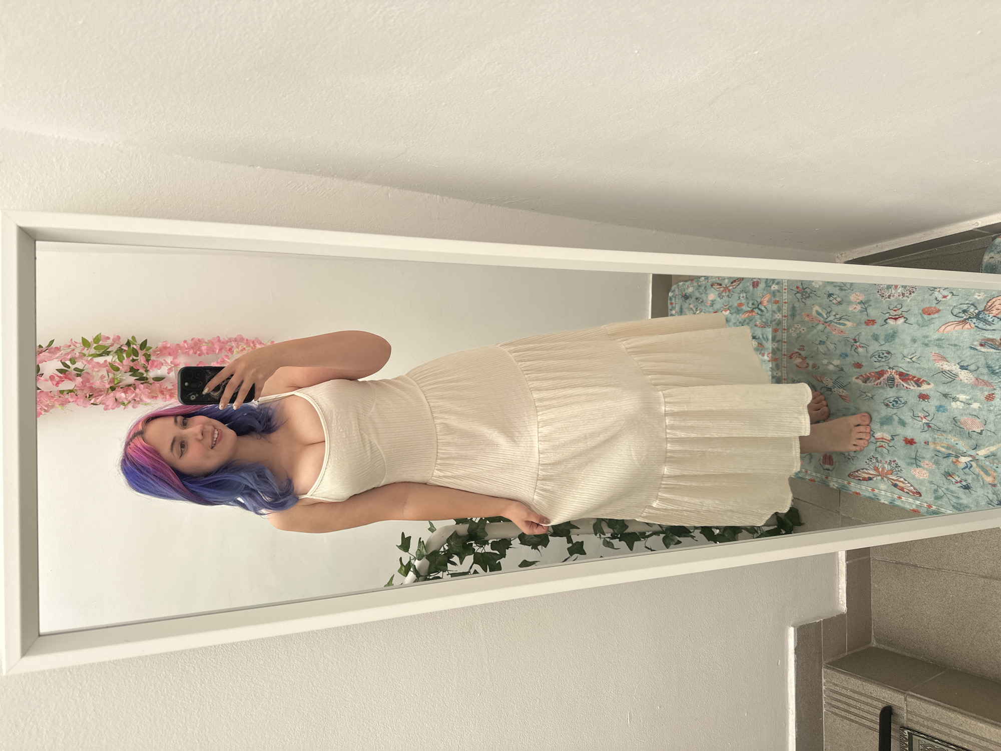 A young woman with a smiling expression is taking a mirror selfie indoors. She has long hair, dyed with vibrant pink and blue/purple streaks, and is wearing a light-colored (off-white or cream) sleeveless maxi dress. The dress features a fitted bodice and a flowing, tiered skirt with a subtle textured or pleated fabric. She holds a dark smartphone in her right hand, visible in the mirror's reflection.
The setting appears to be a brightly lit room, possibly a bedroom or dressing area, with plain white or off-white walls. A large, white-framed full-length mirror reflects the woman and her surroundings. Decorative elements are visible, including pink faux cherry blossom garlands draped on the wall behind her left shoulder and green faux ivy positioned along the bottom edge of the mirror's reflection. On the floor to her right, a light blue or teal rug with a pattern of various insects and butterflies is laid out. She is barefoot. The floor transitions from the rug to light-colored tiles, and further to the bottom right, a step or raised platform with darker gray tiles is visible. There is no discernible text within the image.