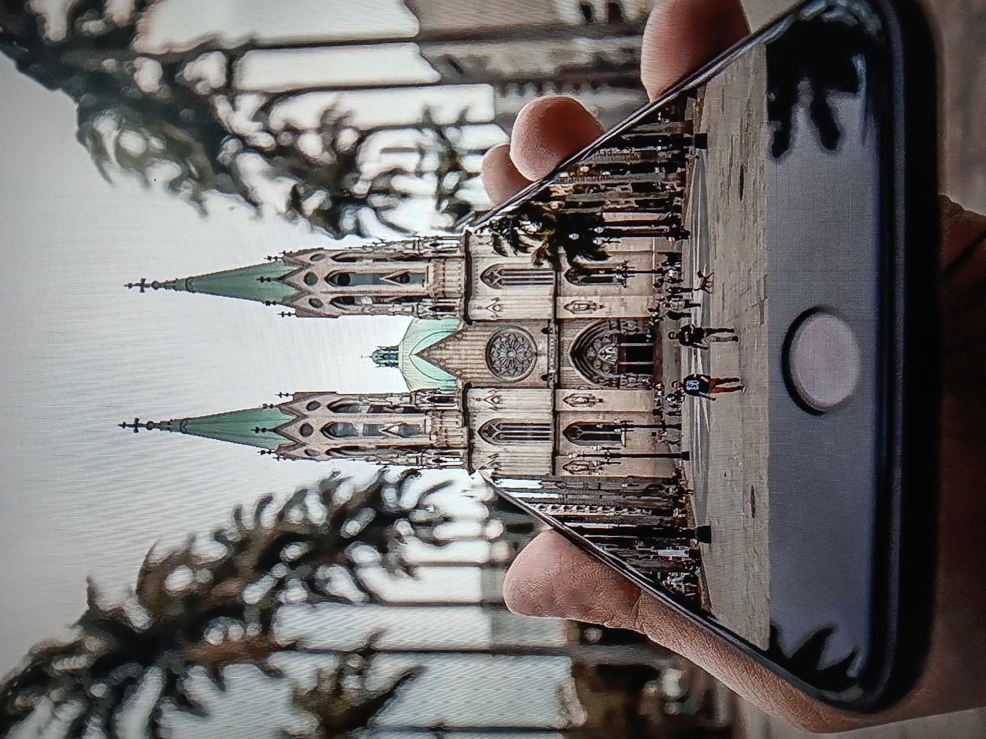 In a medium shot, a hand holds a smartphone, its screen displaying a view of the Metropolitan Cathedral of São Paulo, Brazil. The cathedral's imposing Gothic architecture, characterized by its twin spires and intricate facade, dominates the reflected image. Palm trees line the plaza in front of the cathedral, and people are seen walking and standing in the distance. The hand holding the phone is positioned in the foreground, obscuring the bottom portion of the cathedral's base. The phone's black frame and home button are visible. The scene on the phone's screen suggests a bright, overcast day.