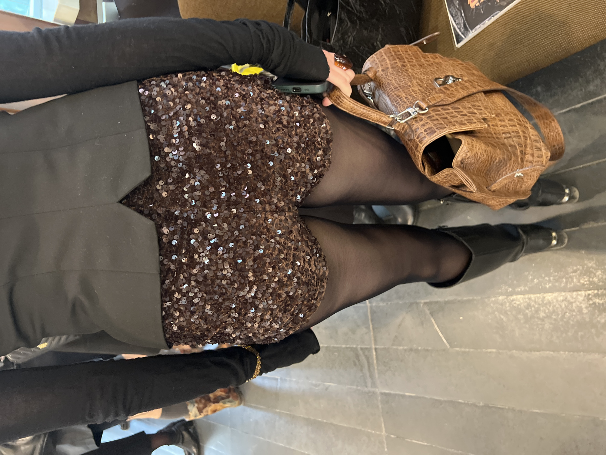 A view from above shows a person seated in an indoor setting. The person is wearing a black long-sleeved top, sequined shorts, black tights, and black boots. Next to the person is a brown, textured handbag. The floor is tiled in grey. A mobile phone is visible on the person's lap. It is not possible to determine the location as Saint Petersburg, Russia.