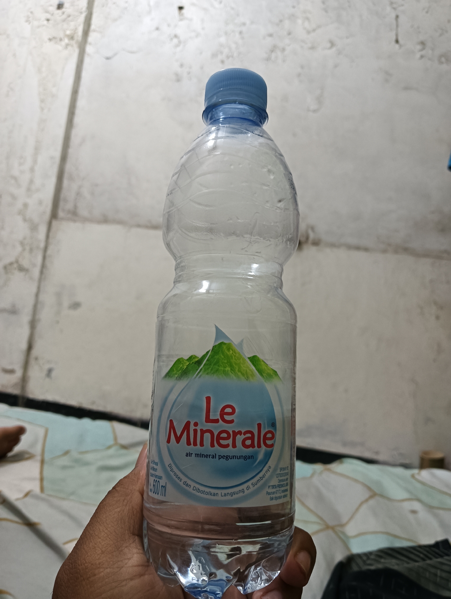 A hand holds a clear plastic bottle of Le Minerale bottled water. The bottle has a blue cap and a label featuring a light blue circle with green mountains and the brand name "Le Minerale" in red text. Below the brand name, it reads "air mineral pegunungan" (mountain mineral water). Text on the label also indicates the volume as "600 ml". The background is a blurry, textured wall and a patterned surface that appears to be a bed. A person's foot is partially visible in the lower left corner of the image. The lighting suggests an indoor setting.
