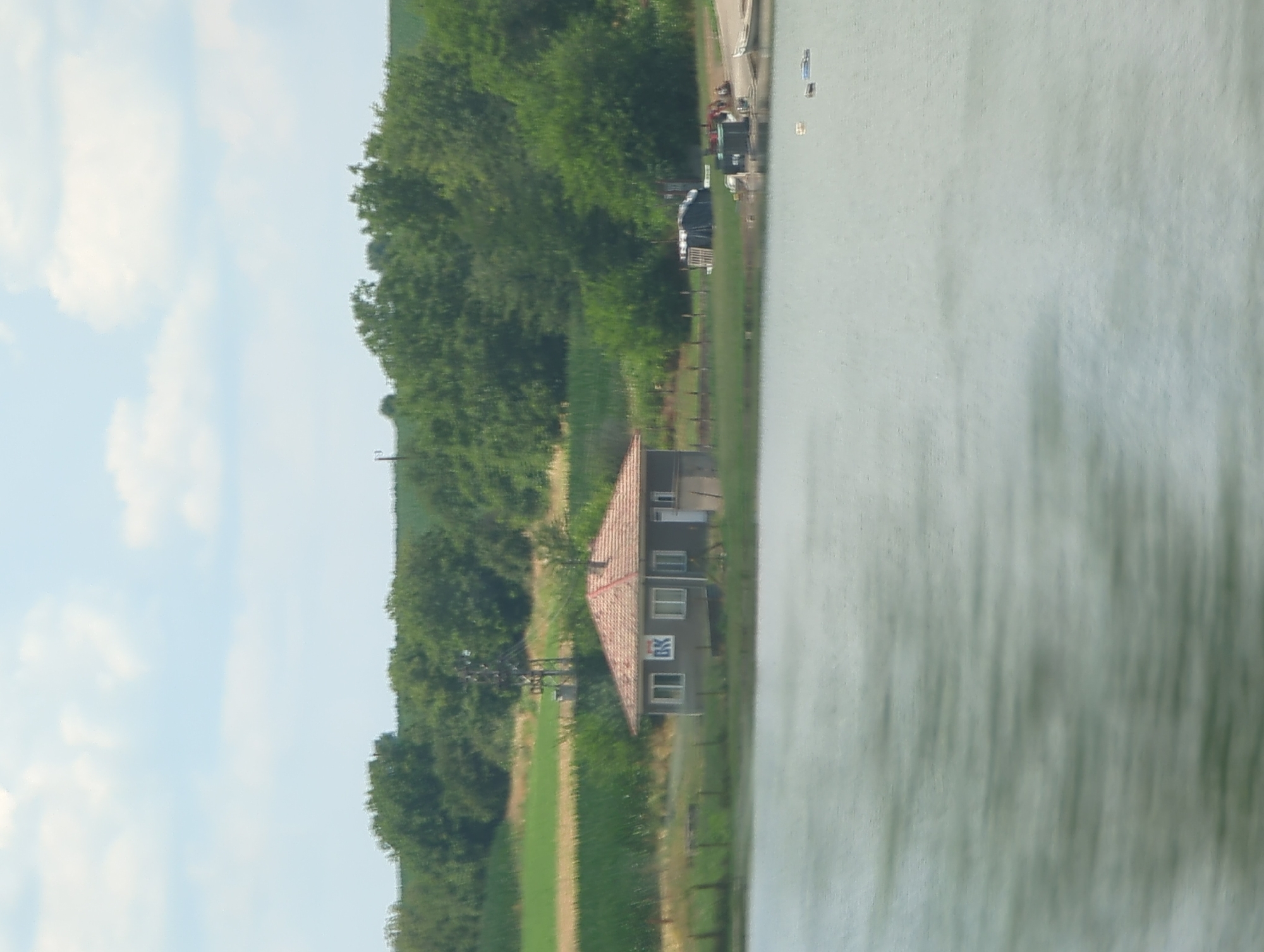 This is a vertically oriented, landscape-oriented photograph taken from a slight elevation. The scene depicts a rural setting in Maslarevo, Bulgaria. In the foreground, a large body of water, likely a lake or river, occupies the right half of the frame, its surface rippled and reflecting the surrounding environment with a muted green hue.
To the left of the water, a narrow strip of land contains a low-slung building with a reddish-brown tiled roof and dark grey walls. The building appears to have several windows and a white door. A prominent logo featuring the letters "BK" in white within a dark rectangular frame is visible on one of the walls. Adjacent to the building, a dark, abstract structure, possibly a piece of equipment or an antenna, stands tall.
Beyond the building, a vibrant green field stretches out, dotted with trees. Further in the background, a dense line of trees forms a dark green band against a bright blue sky scattered with fluffy white clouds. The overall impression is of a peaceful, sunny day in the countryside. There are no people visible in the image. The orientation of the image suggests it may have been taken from a moving vehicle or a high vantage point.