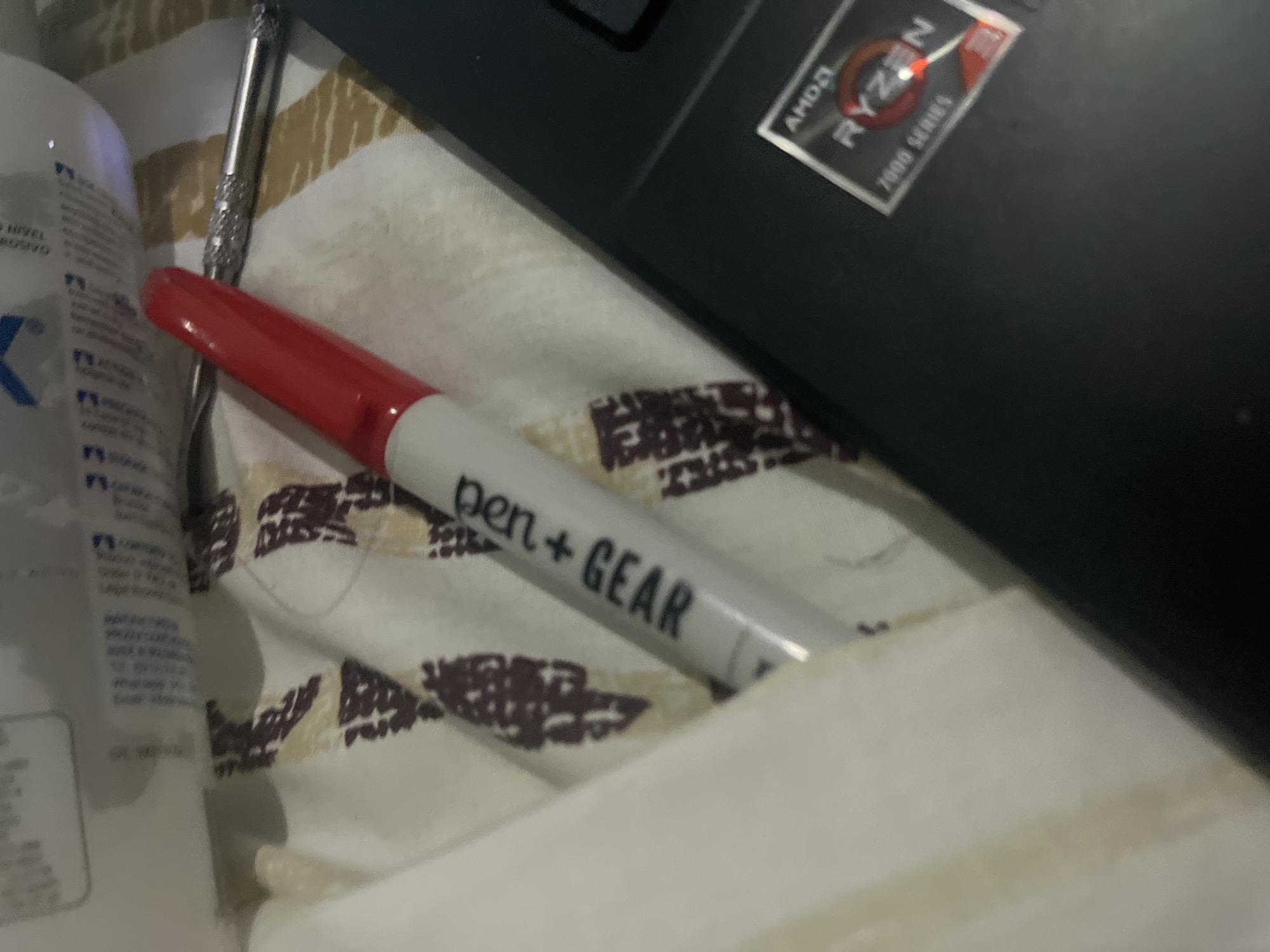 A red and silver marker pen, labeled "pen+GEAR," rests diagonally on a beige and brown patterned fabric, next to a black laptop. The laptop displays an "AMD RYZEN 7000 SERIES" sticker. A small, textured silver object, possibly a tool or pen cap, is situated behind the marker. The background features a blurry white and light beige surface with some faint markings and text. The lighting suggests an indoor setting, possibly a desk or table. No individuals are visible in the image.