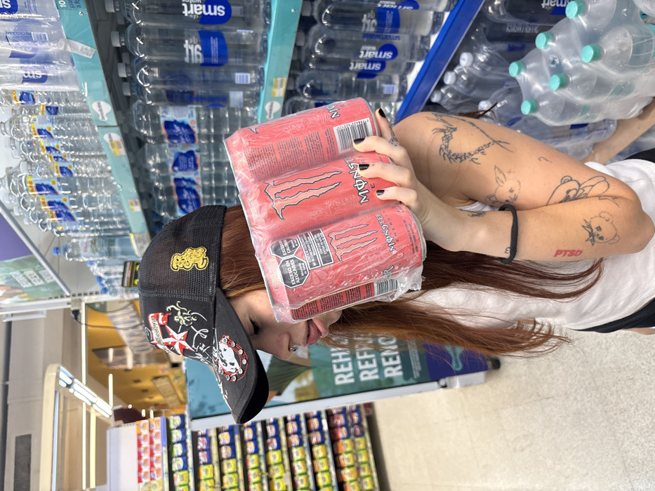 A woman in a black hat with various emblems, including a skull and an octopus, holds a four-pack of Monster energy drinks. Her arms are decorated with tattoos, including depictions of a dragon, a rabbit, and a teddy bear. Text on her arm reads "PTSD." Behind her are shelves stocked with bottled water labeled "Smartwater," as well as shelves filled with various other grocery items. The scene is in a store setting.