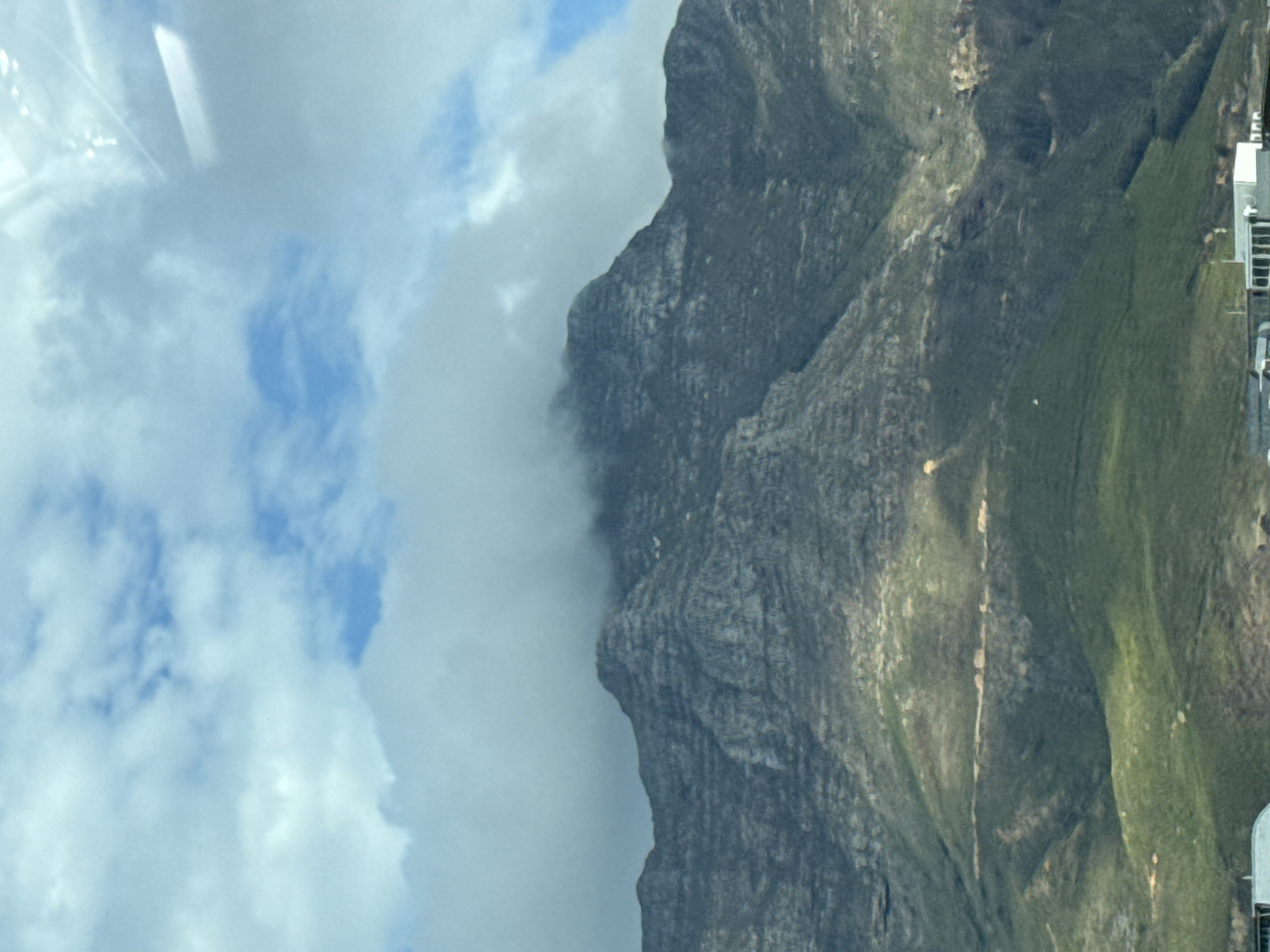 The image depicts a partial view of Table Mountain in Cape Town, South Africa. The mountain's rocky cliffs are visible, with areas covered in green vegetation. A thick layer of clouds obscures part of the mountain. The sky shows blue patches interspersed with white clouds. Buildings are present along the mountainside. A window with a slight reflection is visible.