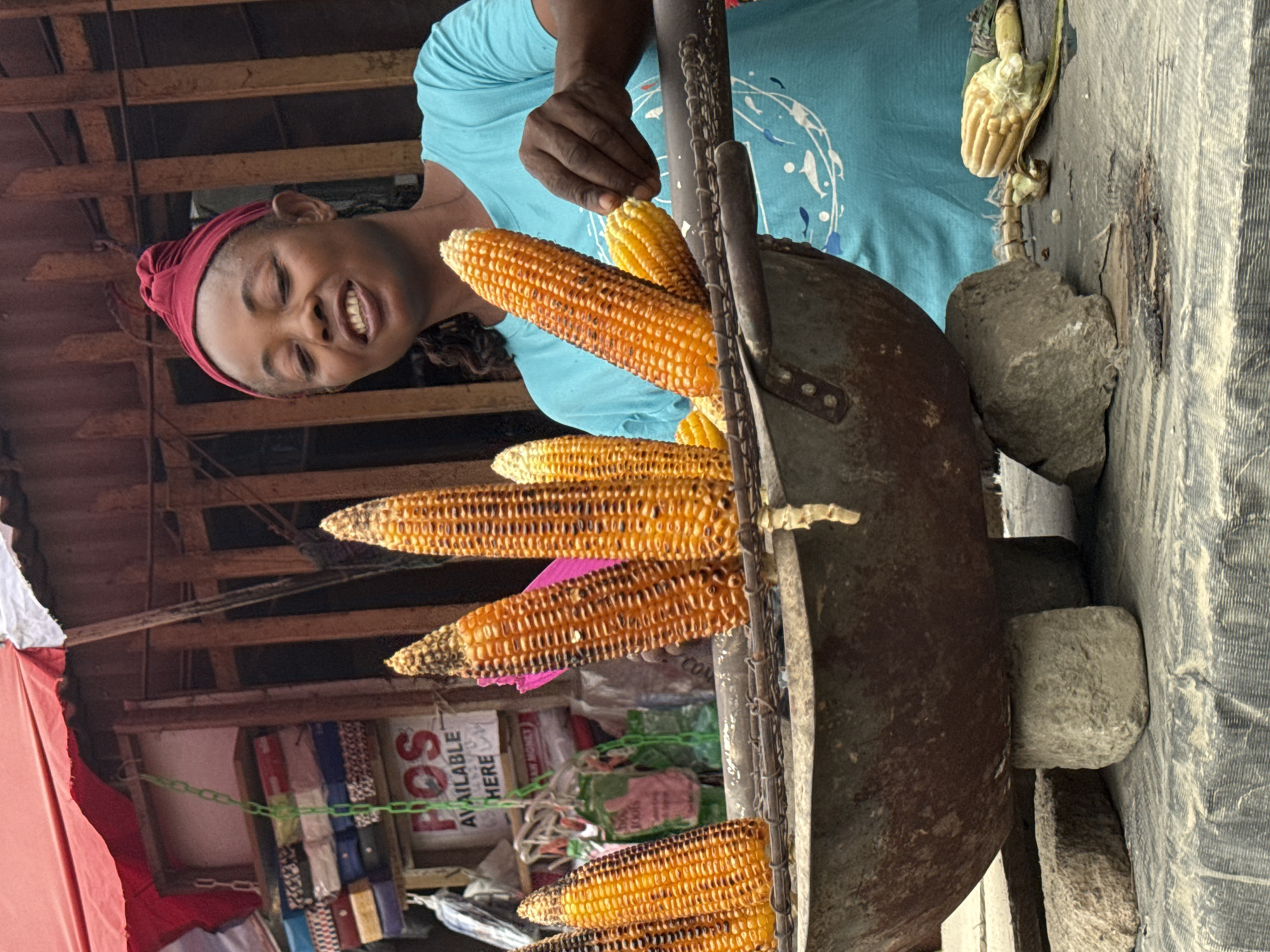 A Nigerian woman, wearing a blue t-shirt and a red headscarf, stands at a street stall in Lekki, Nigeria, preparing to roast corn. She is smiling and looking to her right. Several ears of corn are skewered on a metal rod, which is suspended over a large, rustic cooking pot filled with embers. The pot is propped up by rough-hewn stones. In the background, a stall with various goods, including packaged items and a sign that reads "POS AVAILABLE HERE," is visible. The scene depicts a typical street-side food vendor in an urban setting.