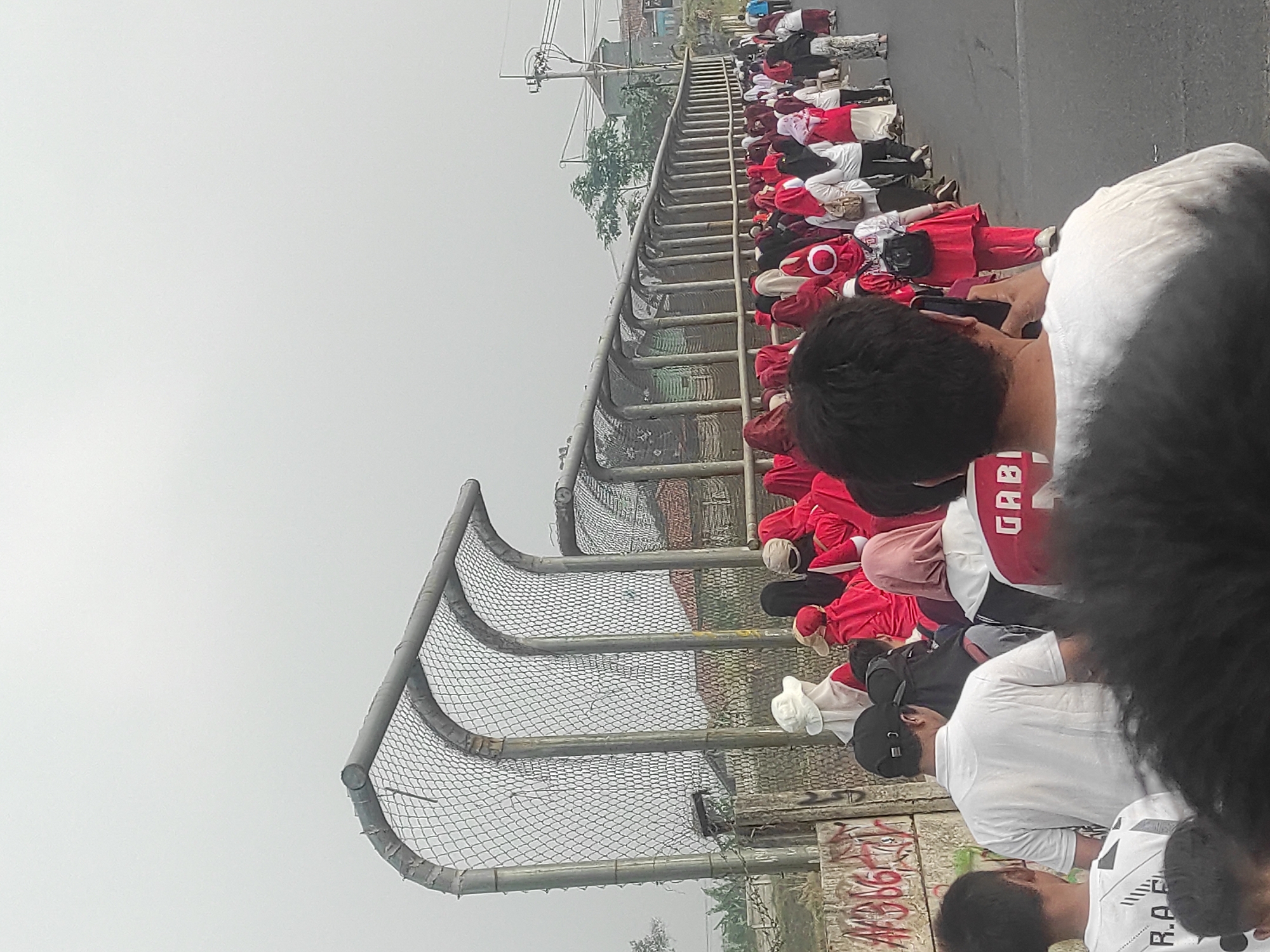 The image shows a group of people, mostly women and girls wearing red headscarves and clothing, walking in a line along a paved road. A metal fence with a mesh covering runs along the left side of the road. In the background, there are houses with red roofs, trees, and a utility pole. The sky is overcast. The people appear to be participating in an outdoor event or procession. The fence has graffiti on its lower section. One person in the foreground is wearing a white shirt and a black baseball cap, while another is wearing a white shirt with "R.A.F." printed on the back. Further down the line, a person is wearing a red outfit and a Santa hat. The setting appears to be a public area in Sumedang, Indonesia, possibly a street or pathway leading to a community gathering.