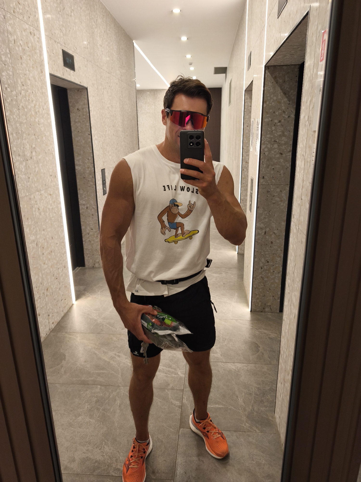 A man stands in a tiled hallway, reflected in a mirror. He is wearing a white sleeveless t-shirt with a graphic of a monkey on a skateboard and the words "SLOW LIFE" printed upside down. He also wears black shorts, a black belt, and bright orange athletic shoes. The man is holding a clear plastic bag containing what appears to be produce and keys in his right hand. He is wearing large, reflective sunglasses with pink lenses. The hallway has a textured, speckled wall and a polished grey tiled floor. Two elevator doors are visible in the background, one on each side of the hallway. The lighting is bright and recessed. The overall setting suggests a modern building, possibly an apartment complex or hotel. The image does not provide any explicit information to confirm the location as Moscow, Russia.