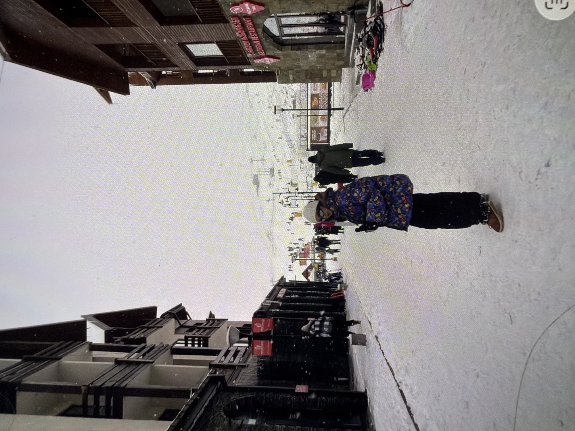 Here is a detailed description of the image:
**Scene:** The image captures an outdoor scene, likely in a ski resort or mountainous area, given the presence of snow and ski lifts in the background.
**Location:** The exact location is uncertain but appears to be a pedestrian area or walkway near buildings. It is in the city of Unknown, Unknown.
**Environment:** The ground is covered with snow. The sky is overcast, suggesting winter weather.
**People:** Several individuals are present in the scene. One person is standing in the foreground, wearing a winter jacket, pants, and a hat. In the background, other people are visible, some walking along the walkway.
**Subjects/Objects:** Buildings are prominent on either side of the walkway. One building on the left has balconies. "Beschen Restaurant" is visible on the building to the right. Ski lifts are seen in the distant background.
**Actions:** The person in the foreground appears to be standing still, while others in the background are walking.
**Interactions:** There are no clear interactions between the people visible in the image. The overall atmosphere is one of a populated but not crowded winter resort area.