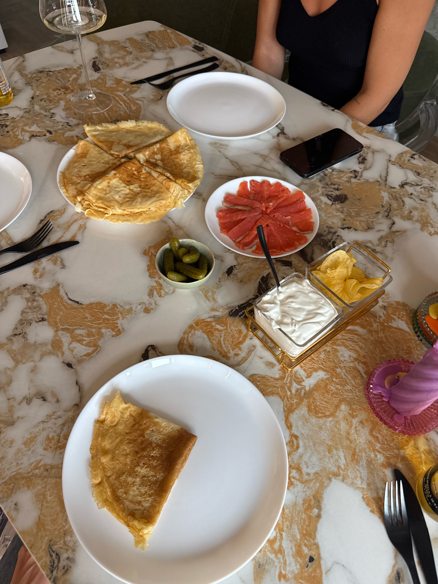 A top-down view of a marble table in Rostov-on-Don, Russia, displays a meal setting with various food items and place settings. The table has a distinctive swirling pattern of beige and brown.
In the foreground, a white plate holds a folded crepe. To its left are black fork and knife. To the right of this plate, a clear plastic container with a gold frame holds two compartments, one filled with a white creamy dip and the other with potato chips. A small black spoon rests in the dip. Adjacent to this container is a small green bowl filled with small pickles.
Further back, a plate of thinly sliced grapefruit is arranged in a circular pattern. Behind the grapefruit is a stack of crepes folded into quarters. A glass of white wine sits to the left of the crepes. To the right of the crepes, a black smartphone is placed on the table.
In the background, a person wearing a black tank top and a silver bracelet is partially visible, seated at the table. A clear plastic chair is also visible. A yellow bottle with an orange label is partially visible on the left side of the table. A pink decorative item with an iridescent sheen sits on the far right. The scene suggests a casual dining or brunch setting.
