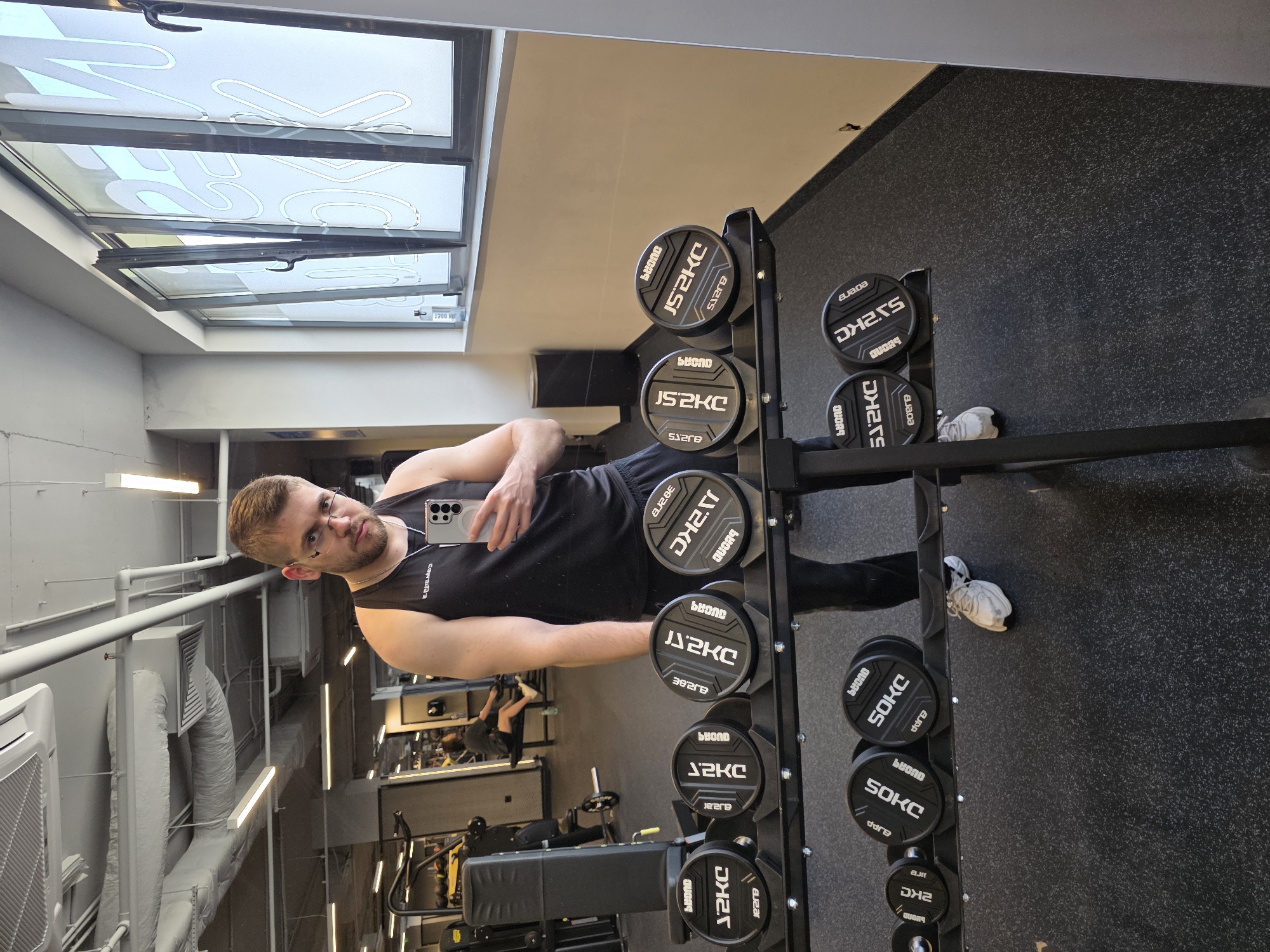 The image displays a selfie taken by a man reflected in a large mirror within a modern gym setting, likely located in Plovdiv, Bulgaria. The man, seen from the chest up, is wearing a black sleeveless top and glasses, with a beard and short hair. He holds a smartphone up, capturing his reflection and the surrounding gym environment.
The mirror reflects a black dumbbell rack positioned to the man's right, laden with various weight plates. These black plates are marked with white lettering, clearly showing different weights such as "DHZ.5KG", "2.5KG", "1.25KG", and "50KG". Many also bear the brand name "beyond" (or its mirrored reflection, "buonD"). The gym floor visible in the reflection is a dark, speckled rubber material.
In the background, beyond the man, the mirror reveals reflections of other gym equipment, including a black exercise bench and additional machines. The ceiling features bright, rectangular light fixtures and extensive, exposed industrial-style ventilation ducts. Above, several skylight windows with white frames and handles are visible, suggesting natural light contributes to the illumination. A numerical marking "1200" is also reflected near the skylights. The overall scene depicts a well-equipped and brightly lit fitness facility.
