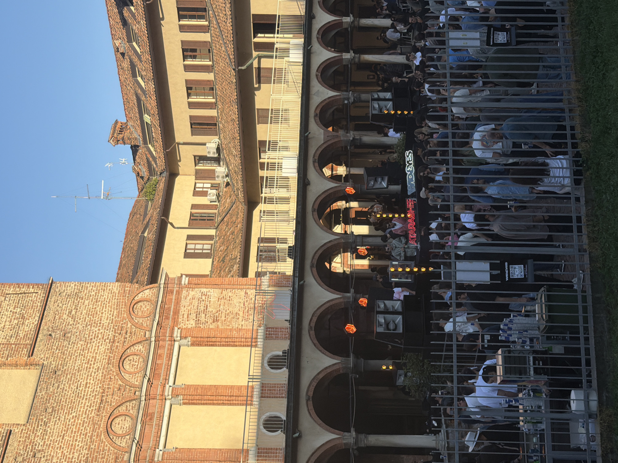 An outdoor scene in Milan, Italy, captures a lively gathering alongside historic architecture under a clear blue sky. The image shows a long, multi-story building on the left, characterized by warm-toned facades, numerous windows with dark shutters, and terracotta roof tiles. Several air conditioning units are visible on its exterior walls, along with an antenna and a chimney pot on the roof. Another building, made of brick with circular decorative elements, is partially visible on the far left.
In the mid-ground, a series of dark arched doorways and columns define an open-air or semi-covered area where a crowd of people is gathered. This area appears to be a venue for a social event, possibly a bar or concert. Within this space, numerous individuals are standing, and some are seated at tables, engaging with each other. Towards the right, a DJ booth or bar setup is visible, featuring large speakers and what appears to be bottles arranged. Artificial lighting, including orange-glowing fixtures, illuminates this darker section, suggesting it might be late afternoon or early evening, or simply a shadowed area contrasting with the sunlit buildings.
The gathering area is partially enclosed by a metal fence or railing, through which many attendees are visible. Notable details include a black sign with a white QR code and the text "WIDGES" visible behind the fence. Other visible text includes "TARASHA" (or "TARASHII") and "SEGS" (or "SEGOS") on signage related to the event or venue. The ground in the foreground is covered with green grass. The overall atmosphere is one of a vibrant urban event.
