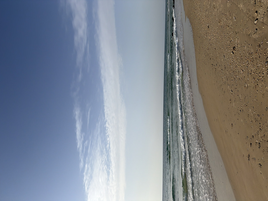 This image captures a wide-angle view of a beach at Beit Yanai, Israel, under daylight conditions. The scene is stratified horizontally into three primary elements: sky, sea, and sand. The upper portion of the frame displays a clear blue sky, with scattered, thin, white cirrus-like cloud formations concentrated across the mid-sky. Below the sky, the Mediterranean Sea occupies the central section, characterized by greenish-blue water with multiple distinct waves breaking towards the shore, producing white foam. The ocean extends to a faint, flat horizon line. The lower portion of the image depicts a light brown sandy beach, curving gently from the lower left towards the right. The sand surface exhibits numerous small seashells and pebbles, particularly in the drier areas, and shows minor indentations consistent with footprints in various locations. A wet line marks the immediate interface between the breaking waves and the sand. No people, animals, or prominent man-made objects are visible in the frame.