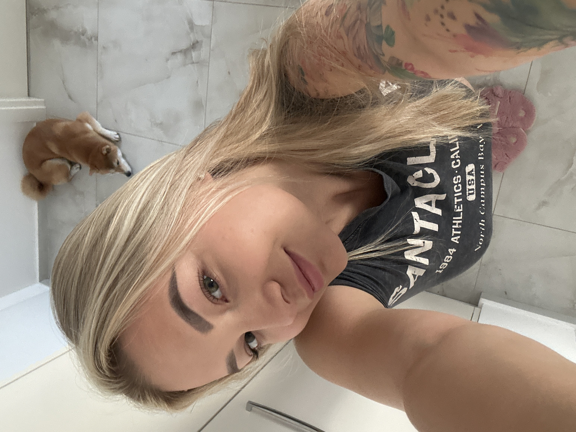 A blonde woman takes a selfie from a high angle, looking down towards the camera with a gentle smile. She has green or hazel eyes, light makeup, and full lips. Her blonde hair falls over her shoulders. She wears a dark t-shirt with text visible on the front, and tattoos cover her left arm and shoulder. The text on her shirt reads "SANTA ATHLETICS CALI 1984 USA North Campus Bay". She is also wearing pink, furry slippers on her feet.
In the background, to the upper left, a small, light brown and white dog, resembling a Shiba Inu, lies curled up on a light-colored tiled floor. The floor appears to be white or light gray marble-patterned tile, extending throughout the visible area. A plain white wall or fixture is visible near the dog and to the far right. The scene is indoors, well-lit, suggesting daytime. The overall impression is casual and relaxed. While the provided context suggests Budva, Montenegro, no specific visual cues within the image confirm this location.