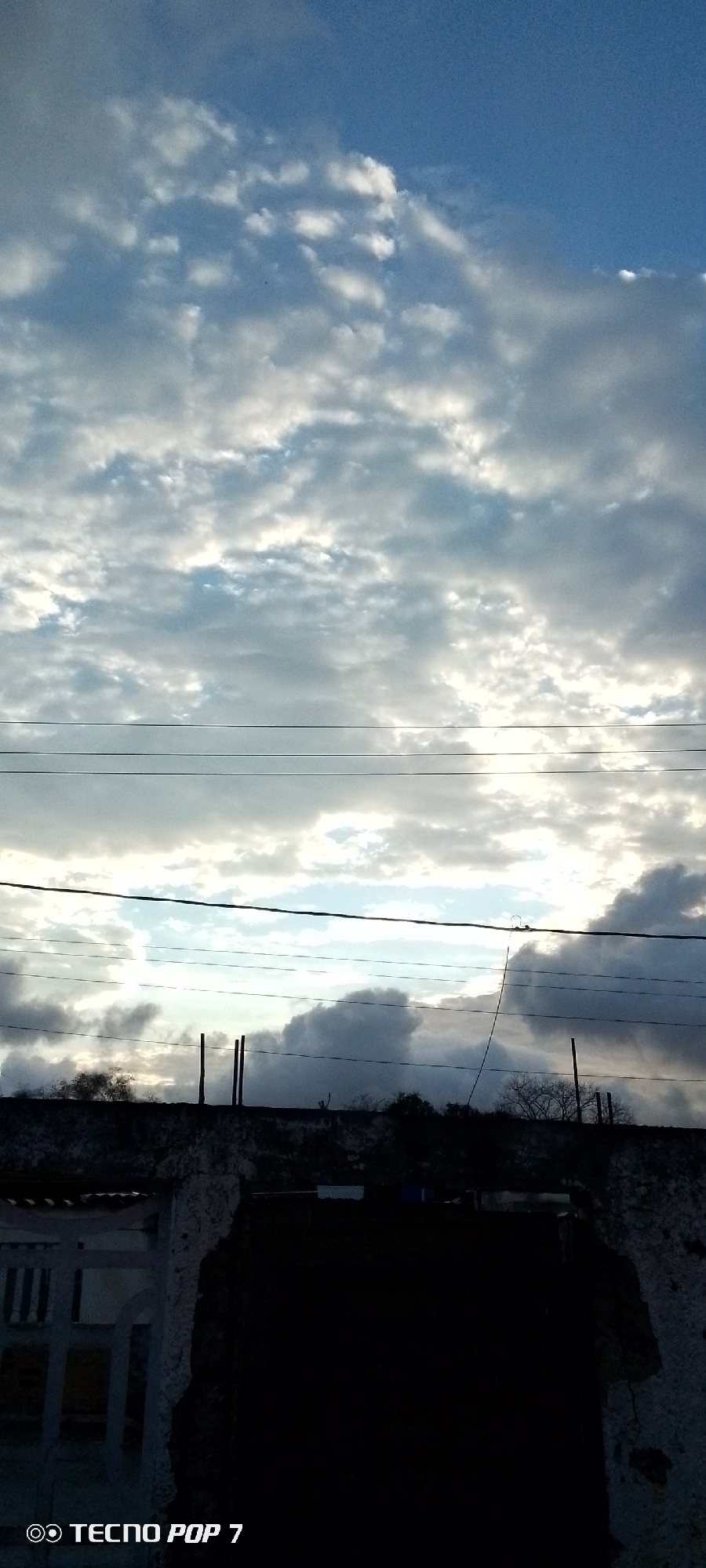 The image shows a view in Altagracia, Venezuela, featuring a sky filled with stratocumulus clouds and electrical wires running horizontally across the frame. Below the wires is a partial view of a rooftop or wall, likely made of concrete or brick, with visible deterioration. Sparse vegetation or trees are silhouetted against the brighter sky in the middle ground. The foreground contains a dark, obscured opening, likely an entrance or window in a building. A portion of a building is visible to the left side. The camera brand "TECNO POP 7" is displayed on the bottom left of the image. The light conditions suggest either dawn or dusk.