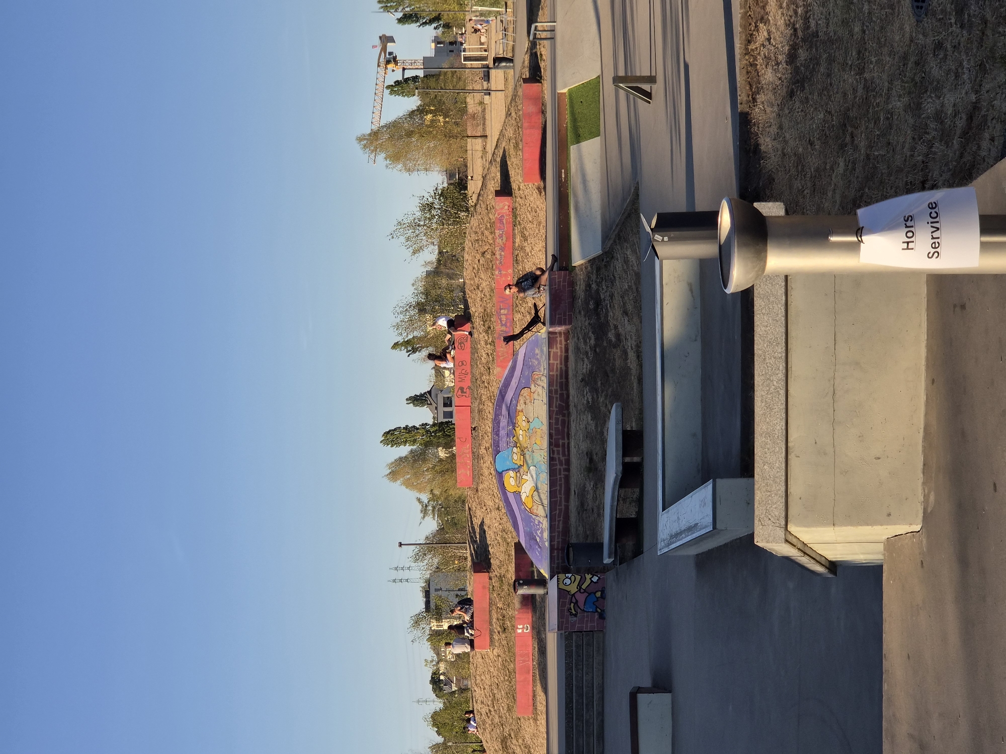The image depicts a public skatepark on a clear, sunny day. The foreground is dominated by concrete structures typical of a skatepark, including ledges, ramps, and a bench. A metallic drinking fountain is visible in the mid-ground, with a white sign hanging from it that reads "Hors Service" (Out of Order). In the background, a grassy embankment is decorated with graffiti, including a depiction of The Simpsons characters. Several people are scattered throughout the skatepark, some sitting on the embankment and others appearing to be observing. Beyond the skatepark, a construction crane and buildings are visible, suggesting an urban or semi-urban environment. The skatepark is located in the city of Sanem, Luxembourg.