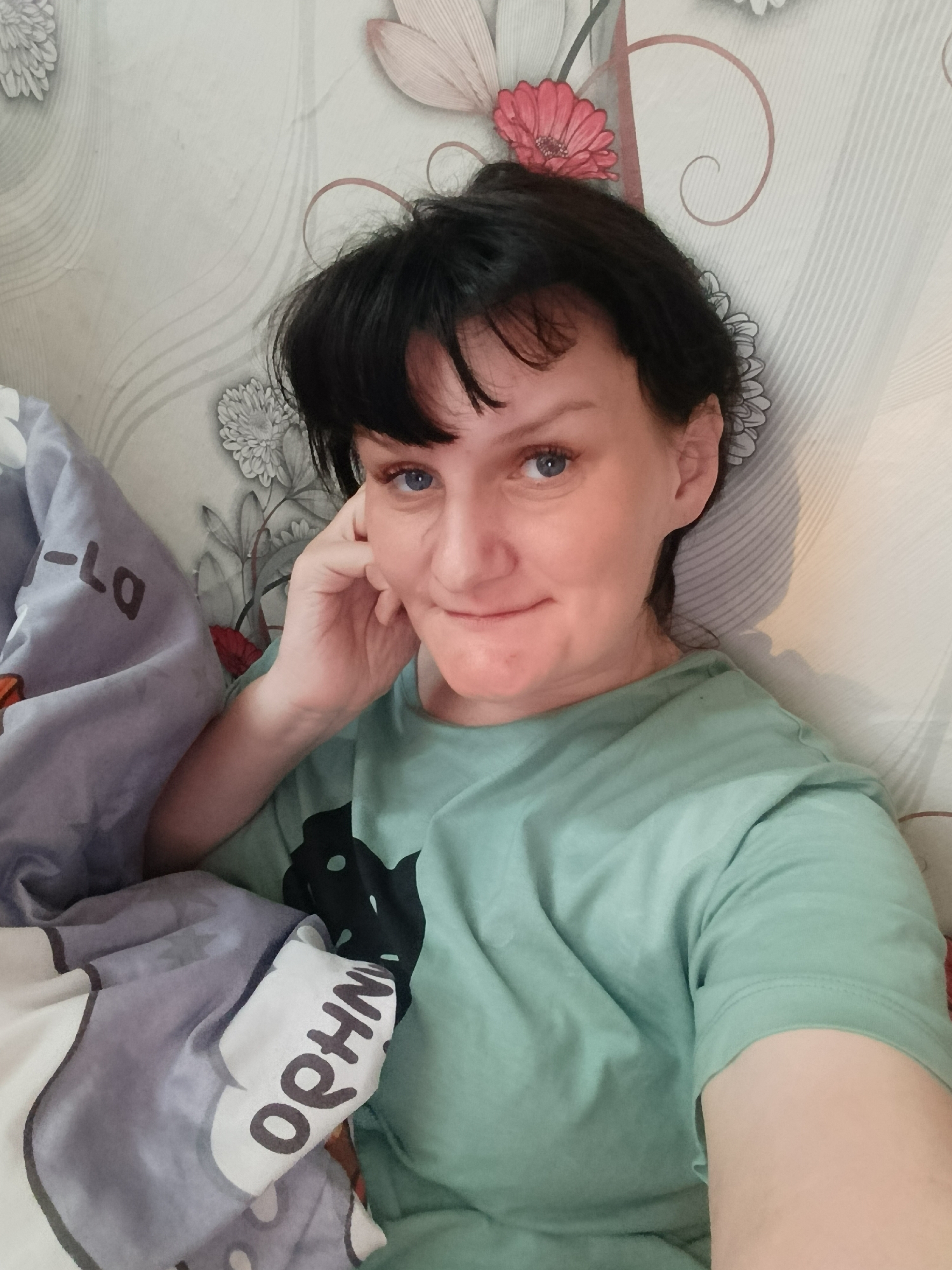 A woman with dark hair and blue eyes is lying in bed. She is wearing a light green T-shirt. Her head is propped on her hand. There is a blanket with cartoon images on it. The wallpaper behind her has a floral pattern. The location is likely an indoor residential space. The image was taken in Фоки, Russia.