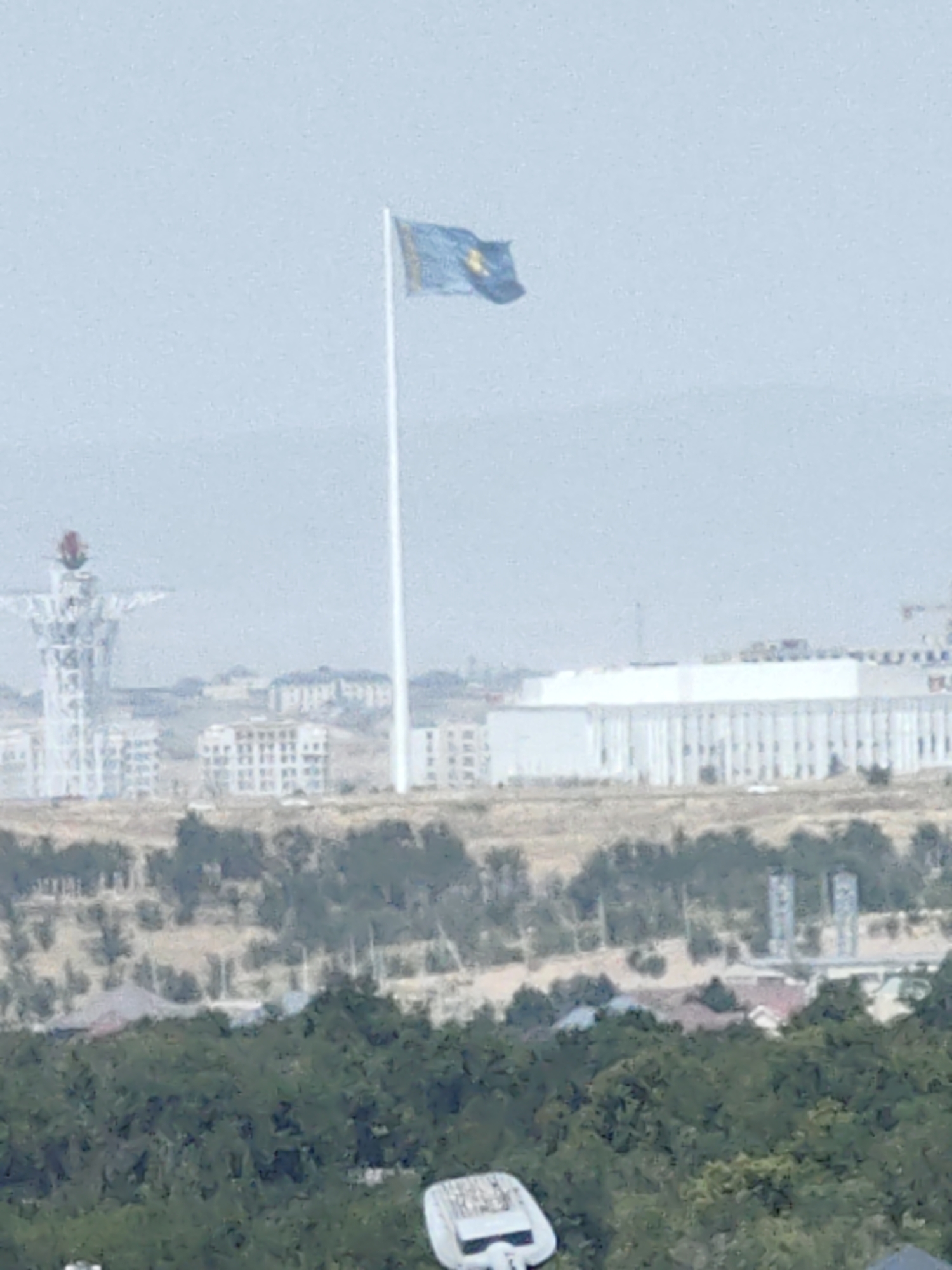 The image captures a wide view of the city of Shymkent, Kazakhstan. Dominating the sky is a very tall flagpole flying a blue flag with a golden sun and eagle. In the background, several buildings are visible, including a large, white, modern structure with many windows and a white tower with a red flame-like top. The landscape between the foreground and the background consists of rolling hills covered with sparse vegetation and some trees. In the lower foreground, a portion of a street lamp fixture is visible. The overall atmosphere is hazy, suggesting a distant or possibly overcast view.