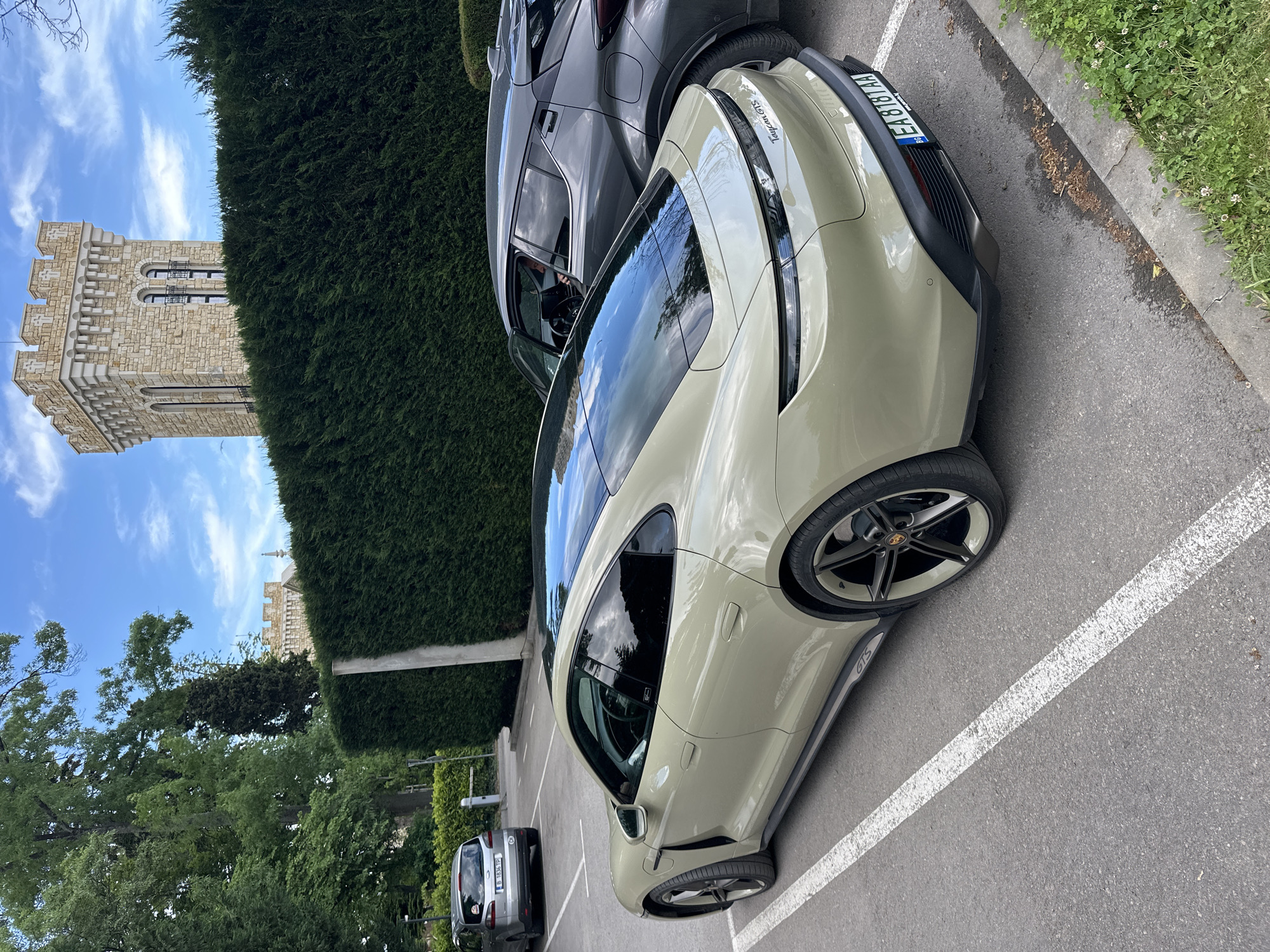 A beige Porsche Taycan GTS is parked in a parking lot adjacent to a tall, manicured hedge. A grey SUV is parked next to it. In the background, a medieval-style stone tower is visible against a blue sky with scattered clouds. The license plate on the Porsche reads "EA 8181 AA". The parking lot is paved, with painted white lines marking parking spaces. The location appears to be near a park or green space in Varna, Bulgaria, given the architectural style of the tower and the vehicle's license plate.