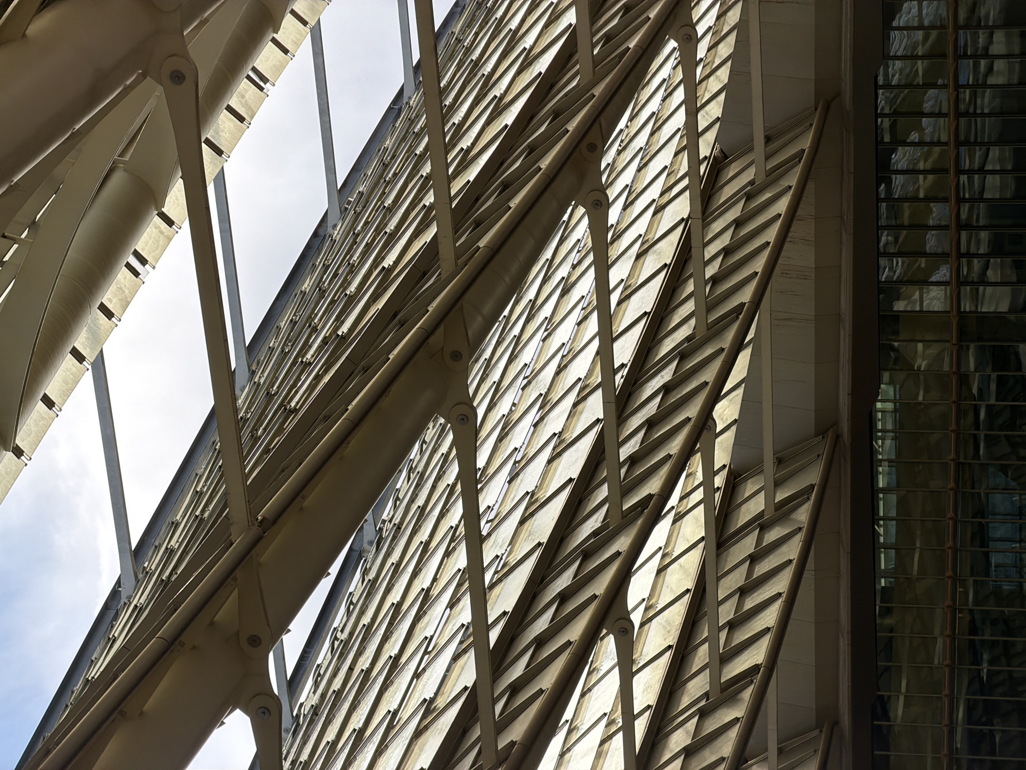 The image displays a close-up, upward-angled view of the distinctive roof structure of the Fondation Louis Vuitton, located in the Bois de Boulogne, Paris, France. The scene focuses on a complex arrangement of overlapping, semi-translucent panels, appearing golden or amber in hue, which form a scale-like pattern across multiple angled and curved surfaces.
Prominent structural elements, including large curved beams and various supporting braces, all painted in a light beige or tan color, are visible, connecting the panel sections. Gaps in the upper roof structure reveal a bright, partly cloudy sky. Below the main roof system, a dark, multi-paned glass facade is visible, reflecting muted, abstract shapes from the interior or surrounding environment. The scene is devoid of people, objects, or any discernible actions or interactions, emphasizing the architectural details of the building's exterior.