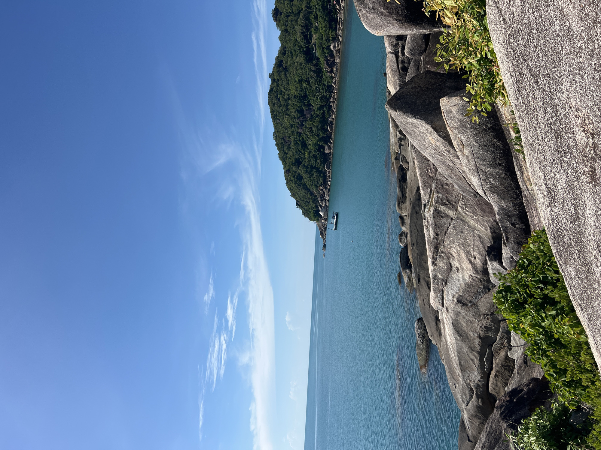 This is a landscape photograph taken from a rocky outcrop overlooking a tropical bay. In the foreground, large, weathered granite boulders are prominent, with patches of vibrant green foliage growing between them. The middle ground features a calm, turquoise sea that stretches to the horizon. A small, white boat is anchored in the bay. In the background, a lush, green, tree-covered hill forms the curve of the bay. The sky is a clear, bright blue with wispy white clouds. The image appears to depict a scenic coastal area, likely a beach or a bay, in the vicinity of Baan Chaweng Noi, Thailand.