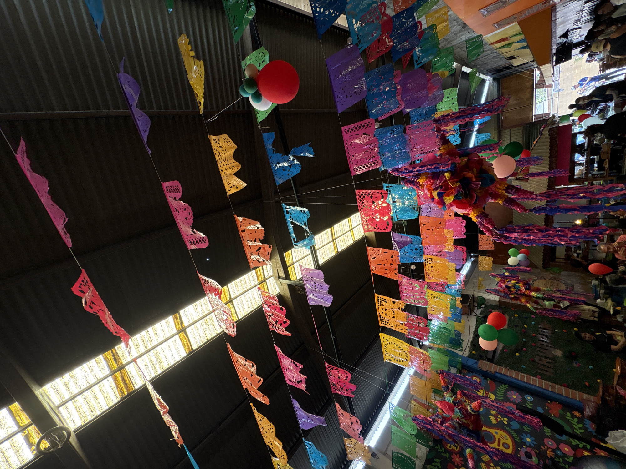 The image shows a view of a decorated indoor space in Ocotepec, Mexico. Numerous colorful papel picado banners hang from the ceiling, creating a festive atmosphere. Balloons are interspersed among the banners. Visible below the decorations is an area with people, flooring and decorated walls. The ceiling is corrugated metal with skylights.