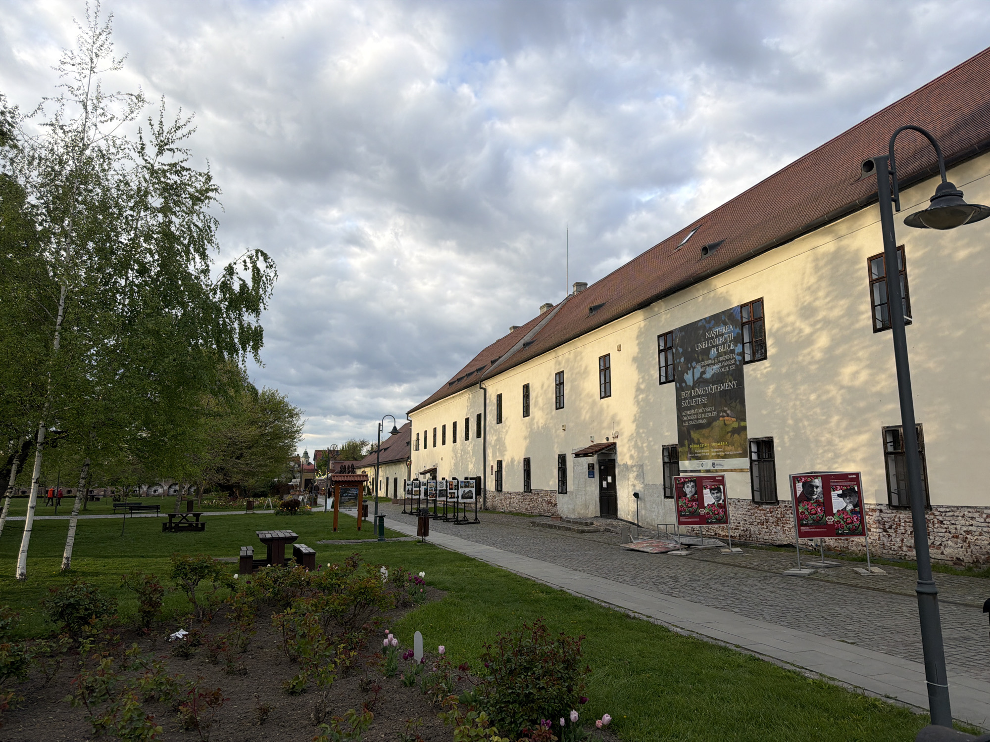 An outdoor wide shot captures a long, cream-colored building with a reddish-brown tiled roof, likely a cultural institution in Târgu Mureș, Romania. The building's facade features numerous dark-framed windows and a large banner advertising an art exhibition. The banner reads "NASTEREA UNEI COLECŢII PUBLICE" and "MOSTENIREA ȘI PREZENŢA ARTER FRANSILVANENE NSECOLUL XXI" in Romanian, with the corresponding Hungarian translation "EGY KÖZGYŰJTEMÉNY SZÜLETÉSE" and "AZ ERDÉLYI MŰVÉSZET ÖRÖKSÉGE ÉS JELENLÉTE A 21. SZÁZADBAN" also visible, along with "GALERIA CETATII" (Citadel Gallery).
Two smaller display posters with portraits and floral designs are positioned on stands outside the building, further indicating an ongoing art exhibit. A paved pathway runs alongside the building, separating it from a vibrant green park area to the left. The park features several birch trees with light bark and fresh green leaves, along with flower beds containing colorful blooms, wooden picnic tables, and benches. A tall, black street lamp stands prominently in the foreground on the right.
The sky is largely overcast with scattered clouds, but hints of sunlight break through, casting a gentle, diffused light on the scene, suggesting either early morning or late afternoon. A few indistinct figures are visible in the distance within the park. The overall atmosphere is peaceful and serene, showcasing a public space dedicated to culture and recreation.