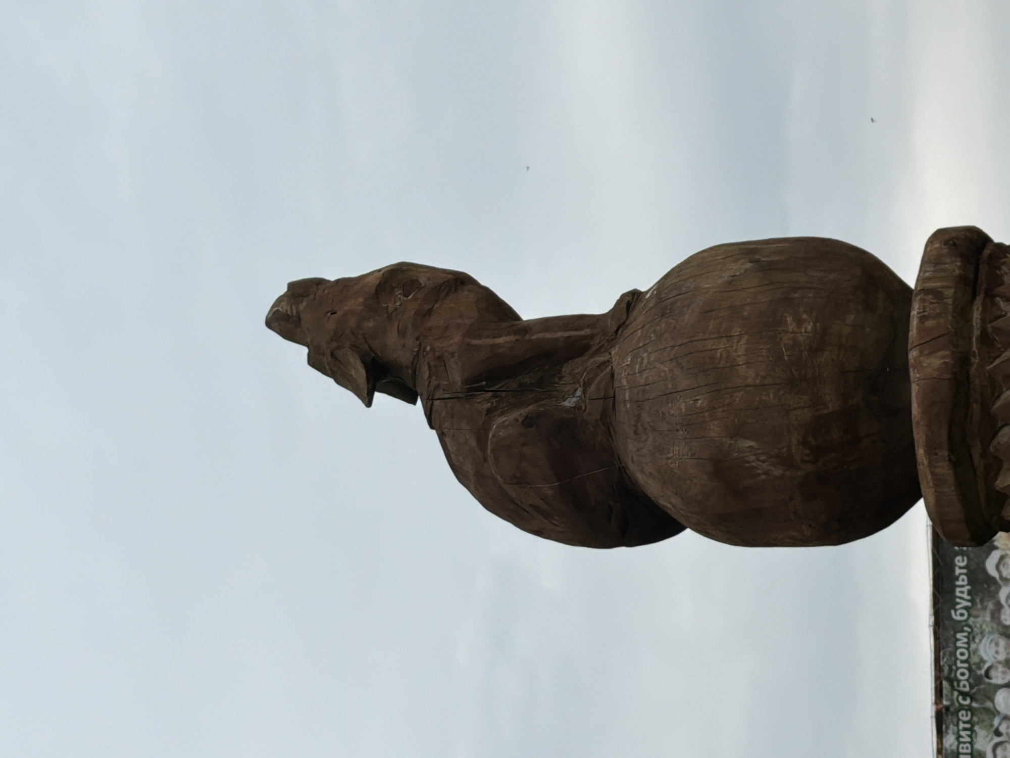 A carved wooden sculpture of a wolf howling stands atop a sphere, positioned against a cloudy sky. The sculpture is dark brown and appears weathered. Below and to the left of the sculpture, a banner with text and images of people is visible. The text on the banner is in Russian and reads "Живите с Богом, будьте!". This sculpture is located in Srednie Kichi, Russia.