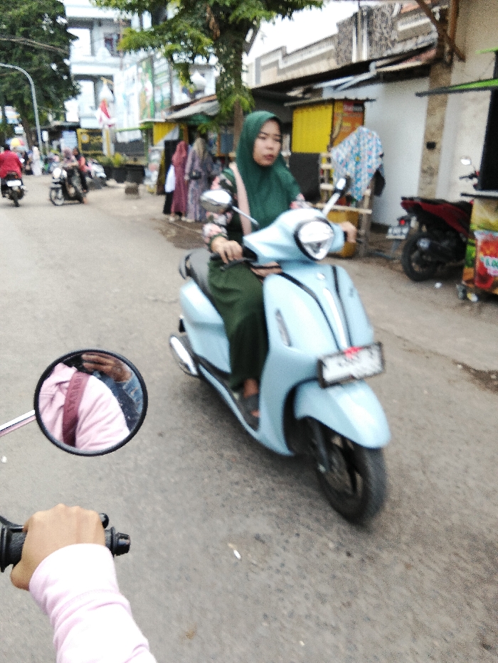 The image depicts an active street scene in Pajarakan Kulon, Indonesia, captured from the perspective of a person riding a scooter. In the foreground, the right hand of a person wearing a light pink long-sleeved garment grips the black handlebar of an unseen scooter. A circular rearview mirror attached to this scooter reflects a partial view of a person wearing a pink top and blue denim.
The primary subject is a woman, approximately 20-30 years old, operating a light blue modern scooter, possibly a Yamaha Grand Filano model. She wears a dark green hijab, a floral long-sleeved top featuring pink, black, and white patterns, and dark green trousers. Her hands are on the handlebars, and she looks forward with a neutral expression. The scooter displays a front license plate, partially obscured.
The scene is set on a paved asphalt road that appears moderately trafficked. In the middle ground, several other individuals on scooters and pedestrians are visible, moving in both directions. On the left side of the street, multiple buildings, including a multi-story structure with a white facade and signage, are visible, interspersed with trees. On the right, a row of small commercial establishments or stalls with brightly colored facades (yellow, green) line the street. Clothing items, possibly traditional garments, hang outside some of these shops. A red scooter is parked near one of these stalls on the right. The overall environment is indicative of a bustling urban or semi-urban commercial area.