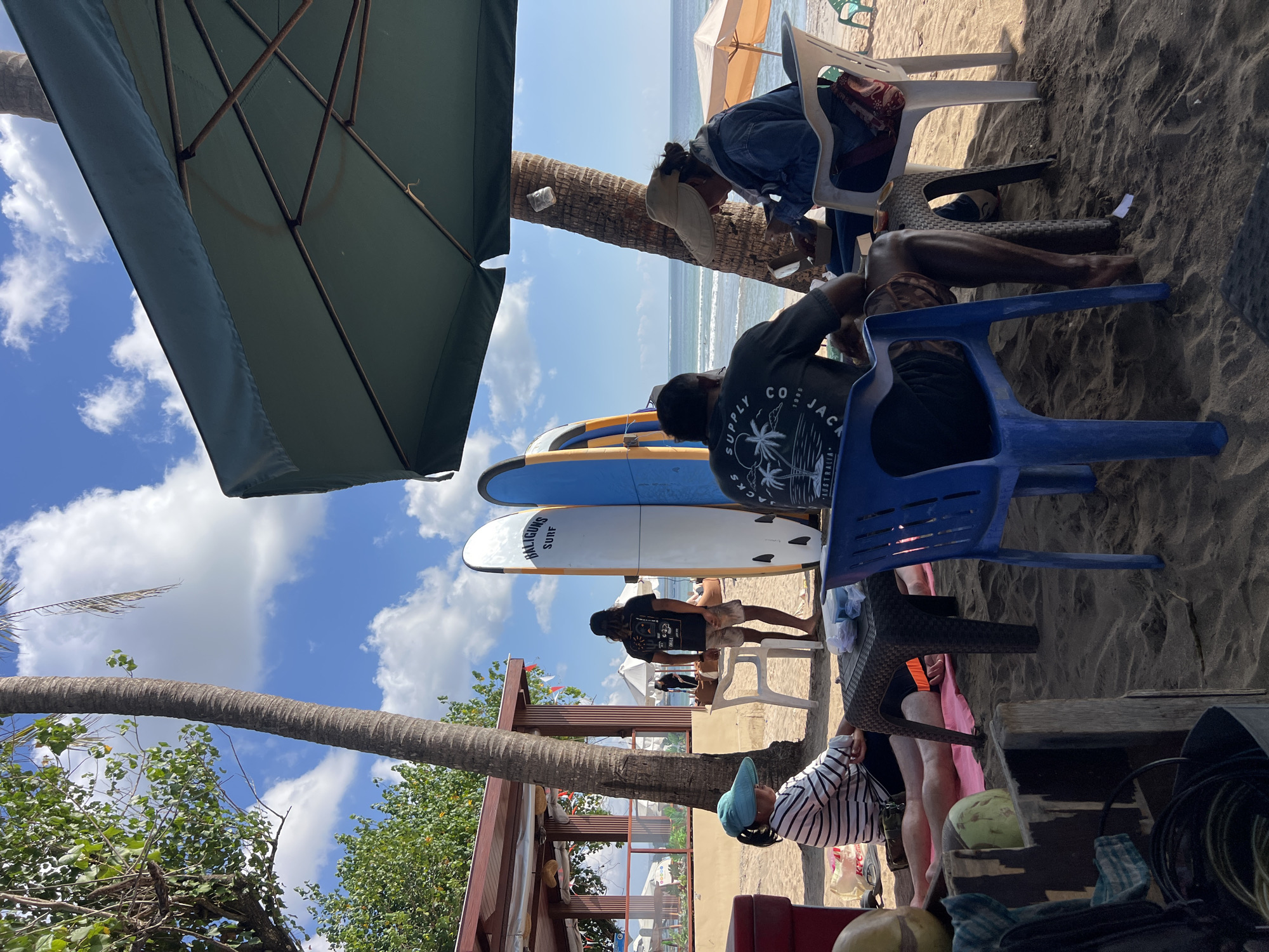 An outdoor scene on a beach in Kuta, Indonesia, depicts a group of people sitting under a large green umbrella. In the foreground, a person wearing a blue and white striped shirt and a light blue sun hat sits on the sand. To their right, a man in a dark t-shirt with a palm tree graphic sits at a dark-colored woven table, facing away from the camera, and appears to be looking at a phone. To the right of the man, a woman wearing a denim jacket and a wide-brimmed hat sits at a white plastic table, also looking at a phone.
Behind them, standing near two surfboards, is a person with long hair, wearing a black t-shirt and shorts, facing away from the camera. The surfboards are white with blue and orange accents, and one has the text "BALIGUNS SURF" printed on it.
In the background, there is a beach structure with a reddish-brown roof and open sides. The ocean is visible beyond the beach, with calm water and a distant horizon. The sand is dark, and palm trees are scattered throughout the scene. Several other umbrellas and chairs are visible in the distance, suggesting a tourist area. The sky is blue with scattered white clouds.