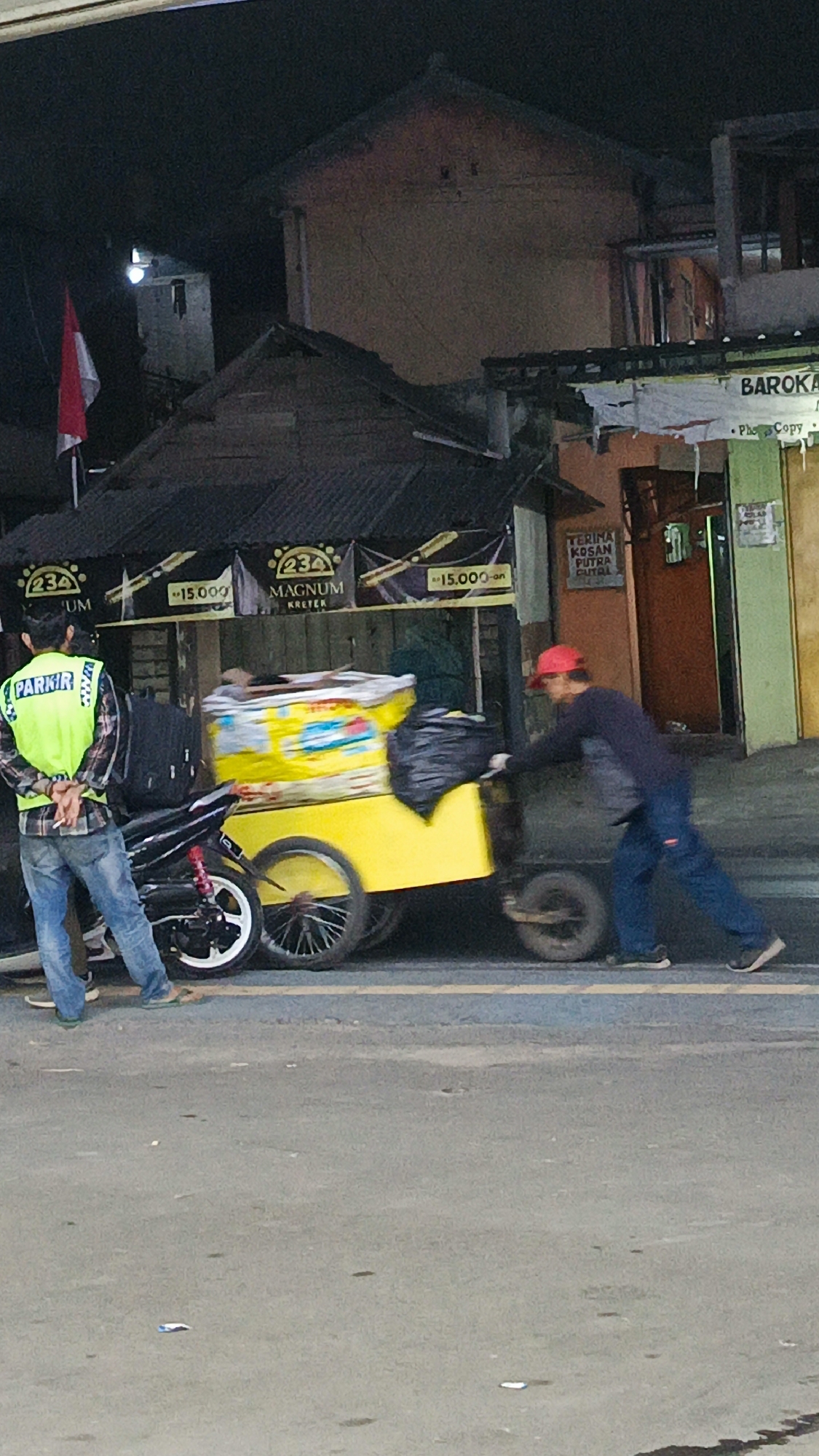 The image depicts a street scene at night in Margajaya, Indonesia. In the foreground, a man wearing a bright yellow vest with the word "PARKIR" printed on it is standing next to a motorcycle. He is wearing blue jeans and appears to be a parking attendant. To his right, a person wearing a red cap and dark clothing is pushing a yellow cart. The cart is laden with goods, including a large black trash bag. In the background, there are buildings, one of which has a sign that reads "BAROKA." A red and white Indonesian flag is also visible on a pole to the left. The overall environment suggests a commercial or residential area operating at night.