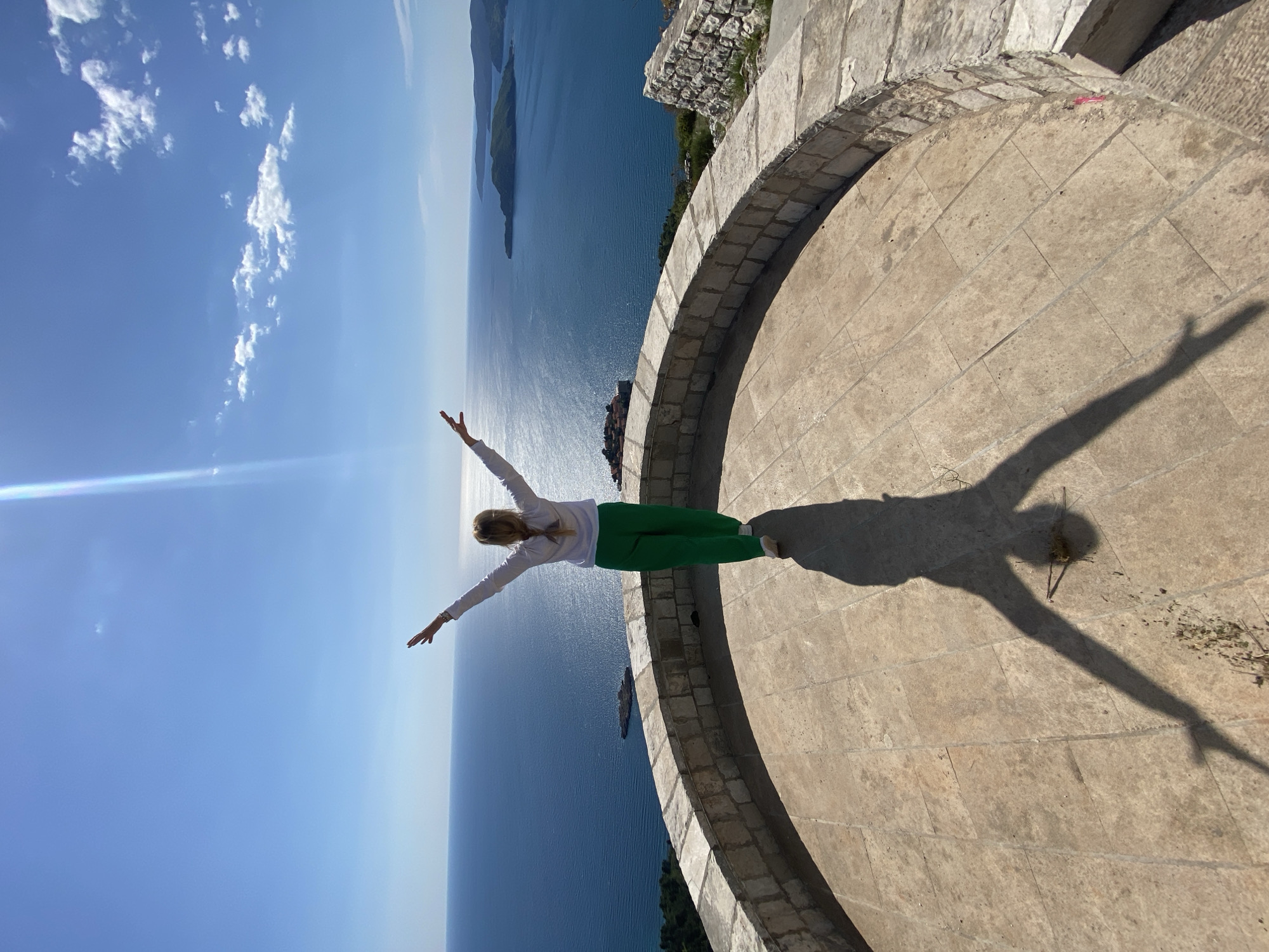 The image, viewed upright, depicts a full-body shot of a person standing on a wide, round stone platform or balcony high above the sea. The person, who appears to be a woman with light-colored hair pulled back, is positioned centrally with her arms outstretched horizontally, a pose suggesting freedom or exhilaration, looking out towards the horizon. She is dressed in a white long-sleeved top and bright green pants.
The platform is constructed from light-colored, rectangular paving stones and features a low, curved stone railing along its edge. A prominent, elongated shadow of the person with outstretched arms is cast on the platform to her right, indicating bright sunlight, likely around midday or early afternoon.
Beyond the platform, a vast expanse of calm, deep blue sea dominates the background, stretching out under a clear, vibrant blue sky dotted with scattered white clouds. Several green-clad islands or landmasses are visible in the distance, enhancing the scenic view. The setting provides a panoramic vista over the water from a high vantage point, characteristic of Sveti Stefan, Montenegro. No visible text is present in the media file.