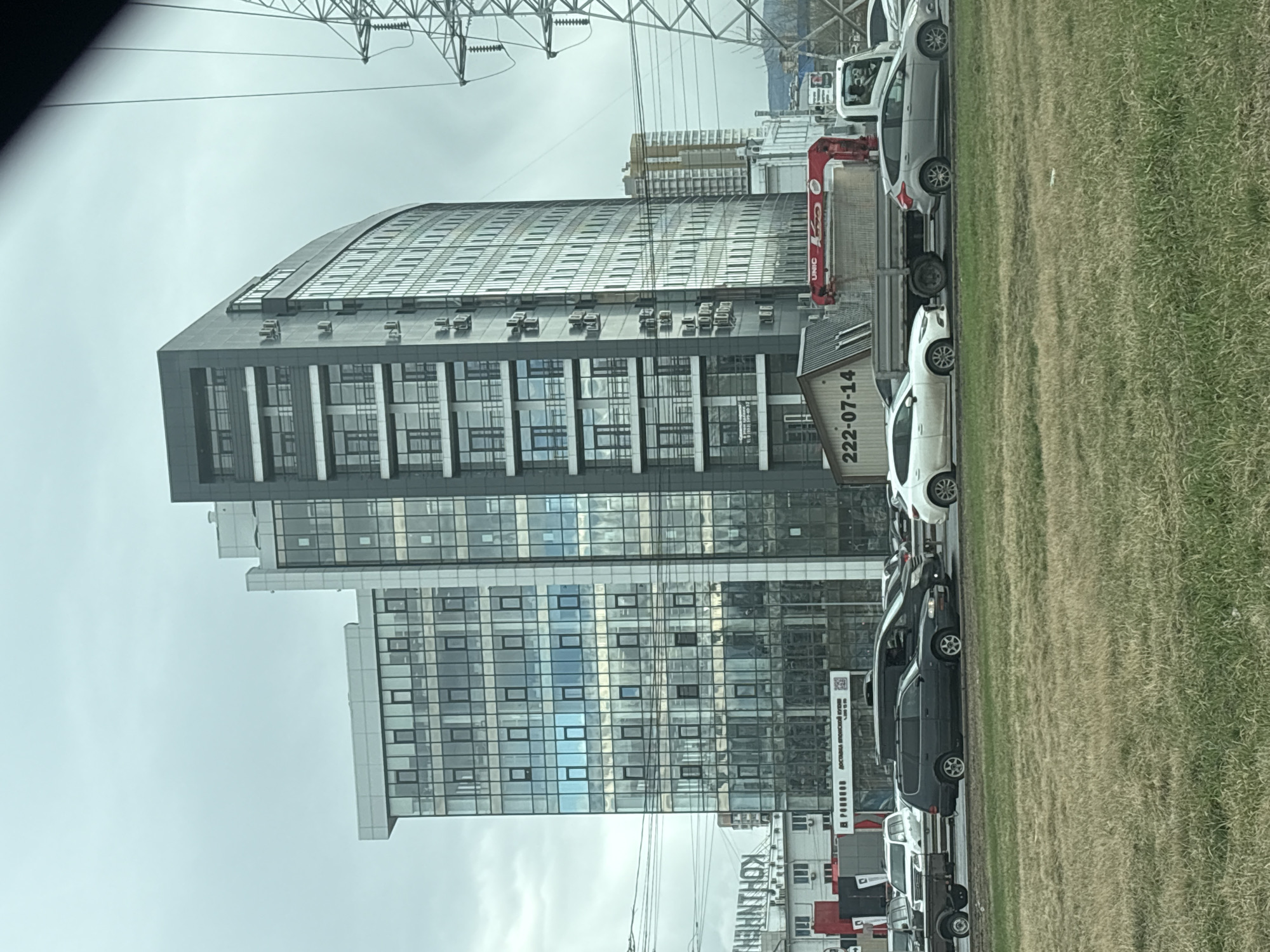 The image captures a view of a multi-story building in Krasnoyarsk, Russia, under an overcast sky. The building features a modern architectural design with a curved roofline on the upper part and a facade predominantly composed of windows and vertical elements. A row of parked vehicles is visible along the edge of a grassy area that runs parallel to the road. Overhead power lines are visible. There is a billboard for "KONZHESTHOY" near the base of the building. A UNIC crane is staged near the building.