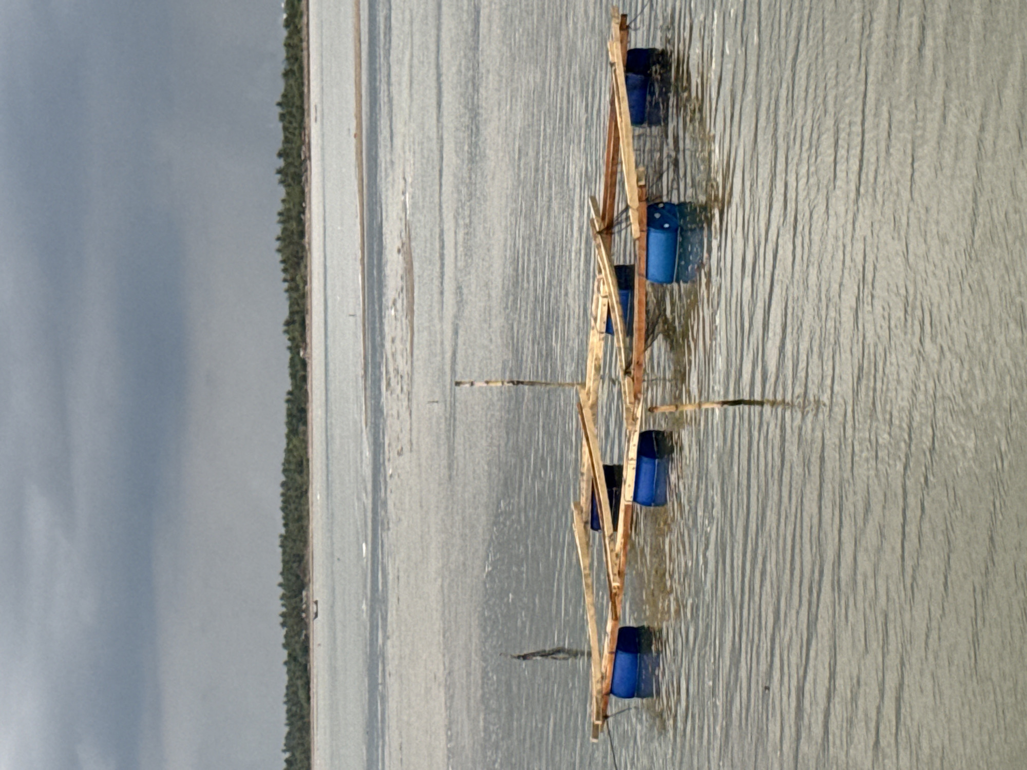 The image shows a man-made wooden structure floating in water. The structure has blue cylindrical containers attached to it, likely for buoyancy. The water has a ripple effect, indicating movement. In the background, the sky is overcast and a strip of land with trees is visible. The location is suggested to be Lhokseumawe, Indonesia.