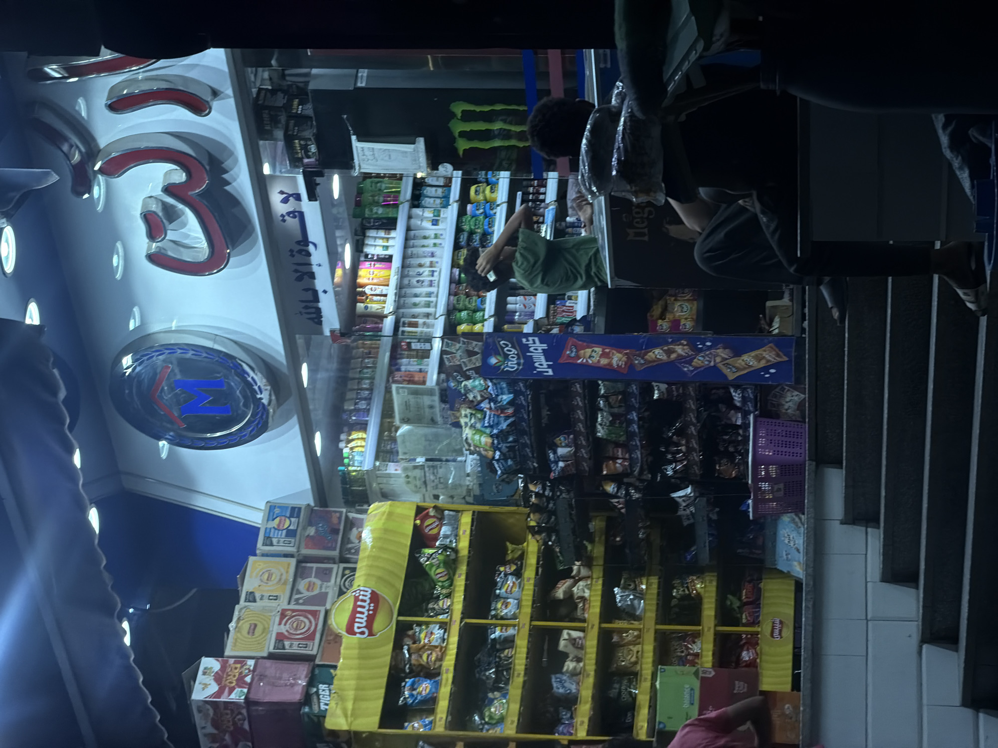 The image depicts the exterior of a small convenience store at night, likely located in the 6th of October city, Egypt. The store is illuminated by artificial lights, highlighting shelves stocked with various products. Prominently displayed on the shelves are rows of snacks, beverages, and what appear to be toiletries or cleaning supplies. A large yellow display unit in the foreground features bags of chips, with Arabic text visible. Above the store entrance, a sign with Arabic lettering and a circular logo with an "M" are illuminated. Two individuals are partially visible. One person, wearing a green shirt, is standing behind a counter inside the store, appearing to be engaged with merchandise. Another person, seen from the back and in darker clothing, is sitting on a low wall or ledge outside the store, to the right of the entrance. The entrance itself has steps leading up to it. The overall scene suggests a typical urban commercial setting at night.