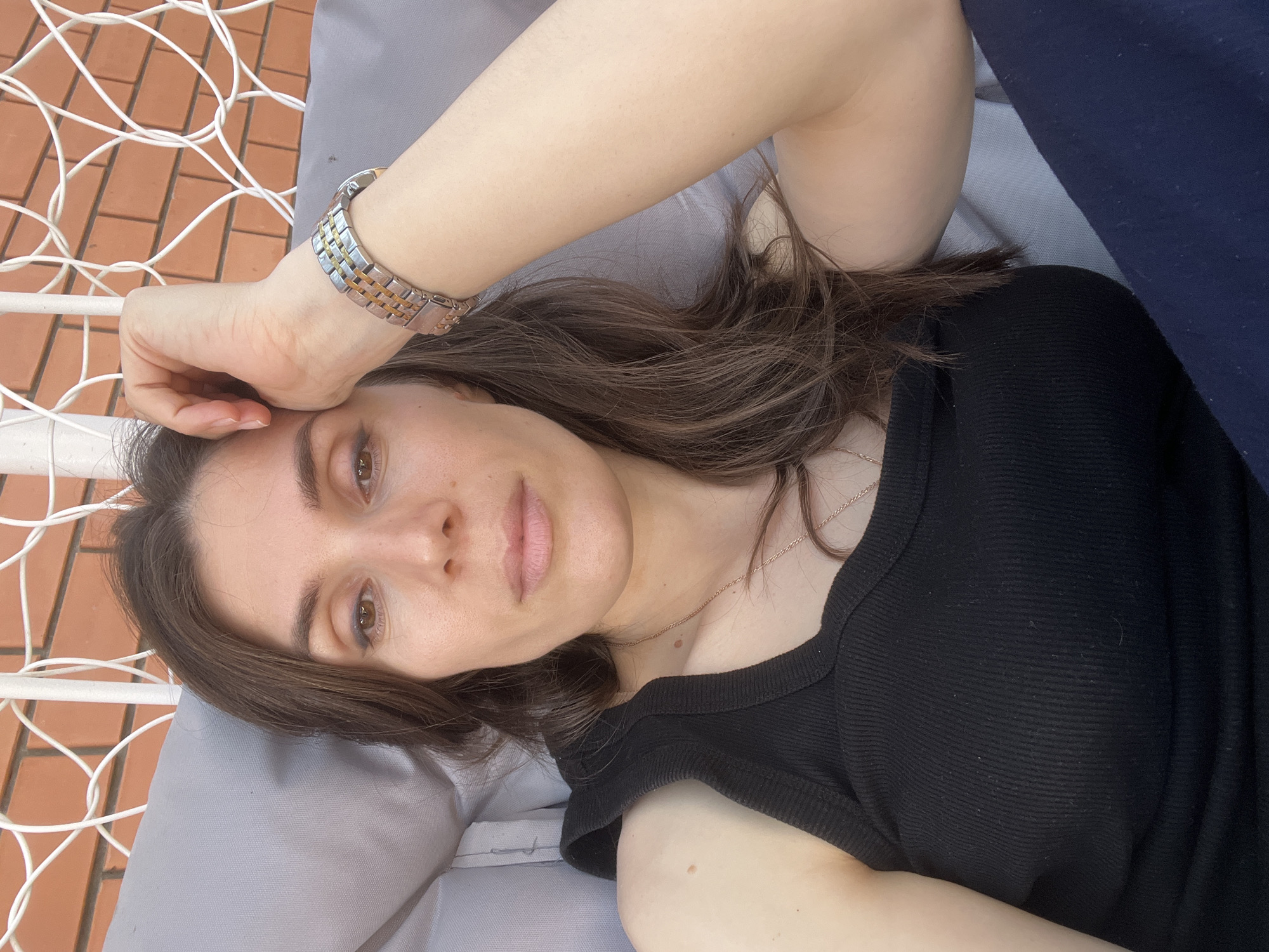 A close-up, high-angle portrait features a woman lying down outdoors, looking directly at the camera. Her head rests on a light gray cushion, with her long, dark brown hair fanned around her shoulders and the cushion. Her left arm is bent, and her hand is placed near her forehead. She has brown eyes and a neutral, calm expression.
She is wearing a black ribbed short-sleeved top. A thin, gold-toned necklace with a small pendant is visible around her neck. On her left wrist, she wears a two-toned metal watch featuring both silver and gold or bronze links.
The background indicates an outdoor setting. To the left, a white metal grid structure, possibly part of a hammock or an outdoor bed frame, is prominent. Below this, an orange or terracotta-colored tiled surface, resembling brickwork, is visible. A dark blue fabric or surface occupies the right side of the frame. The lighting suggests it is daytime. The scene depicts a relaxed moment in an outdoor area, identified as Kaspiysk, Russia. There is no visible text in the image.