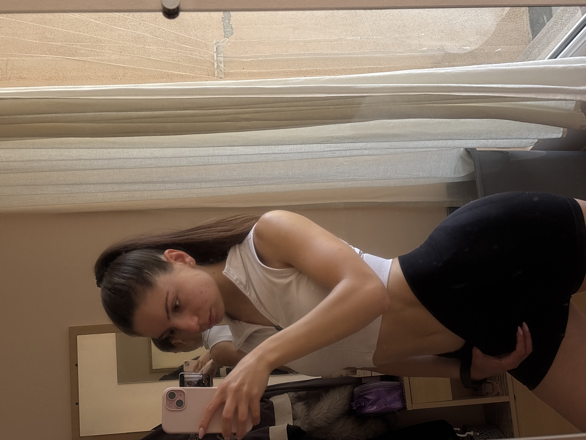 A young female is depicted taking a mirror selfie indoors. She has long, dark hair pulled into a high ponytail and is wearing a white, sleeveless, cropped top with a partial zipper and black athletic shorts. Her left hand holds a light pink smartphone, which she is using to capture her reflection in the mirror. Her right hand rests on her upper thigh or hip, slightly adjusting her shorts. She gazes intently towards the phone's screen.
The indoor environment features light-colored, plain walls. To the left, a rectangular, unframed mirror is mounted, reflecting a portion of the room behind the subject, including a second mirror frame and hanging garments, one with visible fur trim. Below these, a light-toned wooden cabinet is partially visible, topped with a purple, glossy handbag or pouch.
To the right of the subject, a large window is present, covered by vertical, light-colored sheer blinds or curtains. Beyond the window, an outdoor scene is discernible, featuring a textured, light-brown building facade, possibly a balcony or exterior wall. White lines, consistent with a clothesline, are strung across the visible exterior, with a white mounting bracket affixed to the wall. A dark grey object, possibly a waste bin or outdoor furniture, is partially visible in the lower right beside the window. The lighting suggests natural daylight. The user states the location is Varna, Bulgaria.