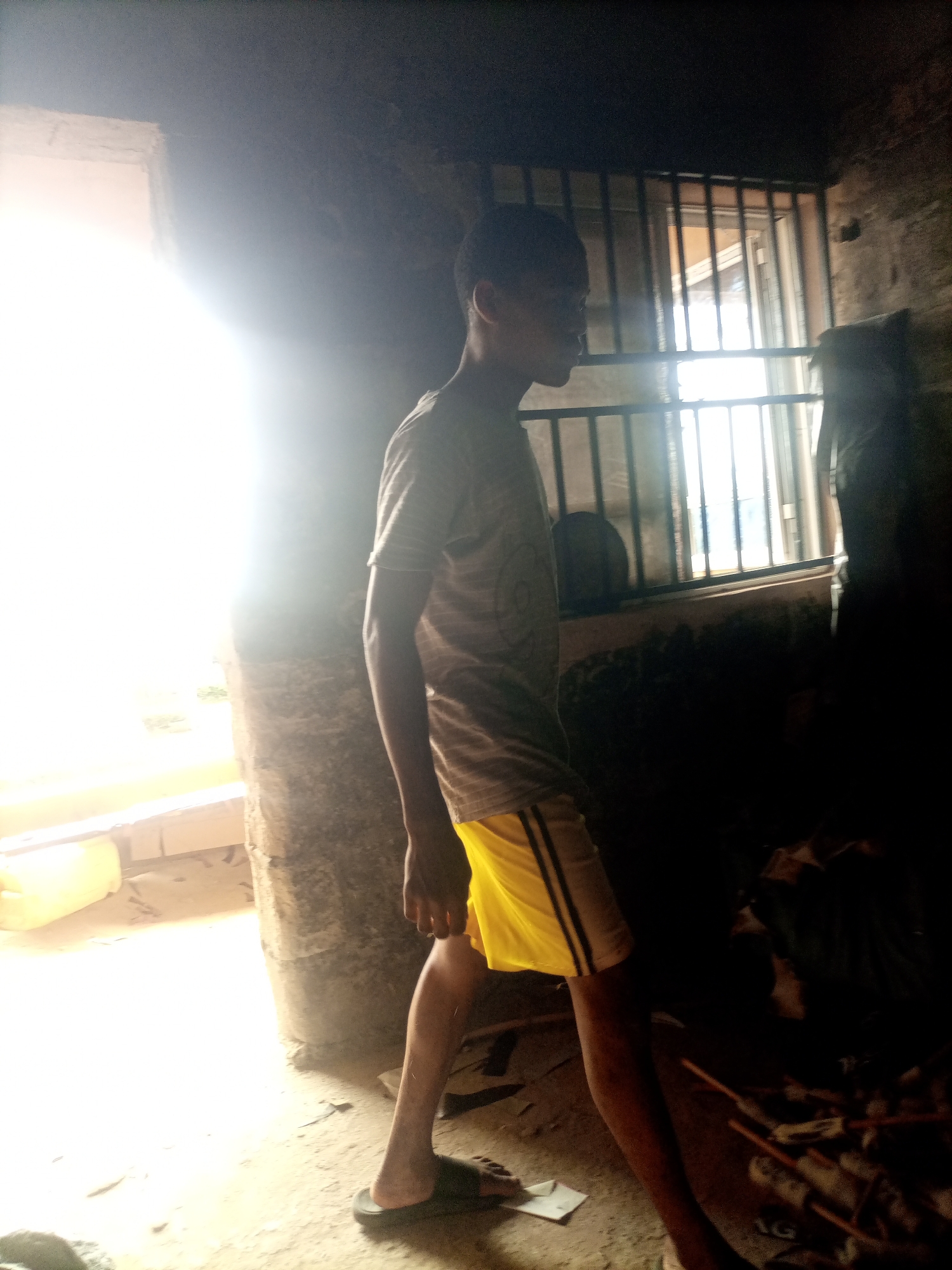 A dark, dimly lit room in Nkpor, Nigeria, contains a young male subject. The subject is wearing a striped t-shirt and yellow shorts. He is standing in profile, facing left, and appears to be walking. The room has rough, textured walls and a concrete floor. A barred window is visible behind the subject, through which daylight is filtering in, creating a bright glare. There is a structural column to the left of the subject and a pile of items on the floor to the right, which are obscured by shadows. The overall lighting suggests an interior space with a strong light source from an opening to the left.