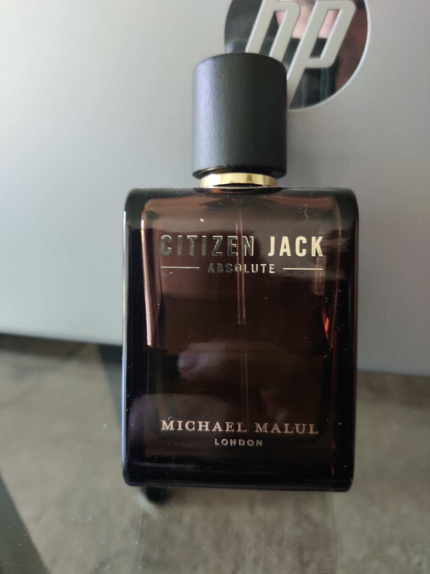 A dark brown, rectangular glass bottle of "CITIZEN JACK ABSOLUTE MICHAEL MALUL LONDON" perfume is centered in the foreground, resting on a glass surface. The bottle has a black cap with a gold band. Behind the bottle, a silver laptop with an HP logo is visible, slightly out of focus. The background appears to be a wall. The location is not specified in the image.