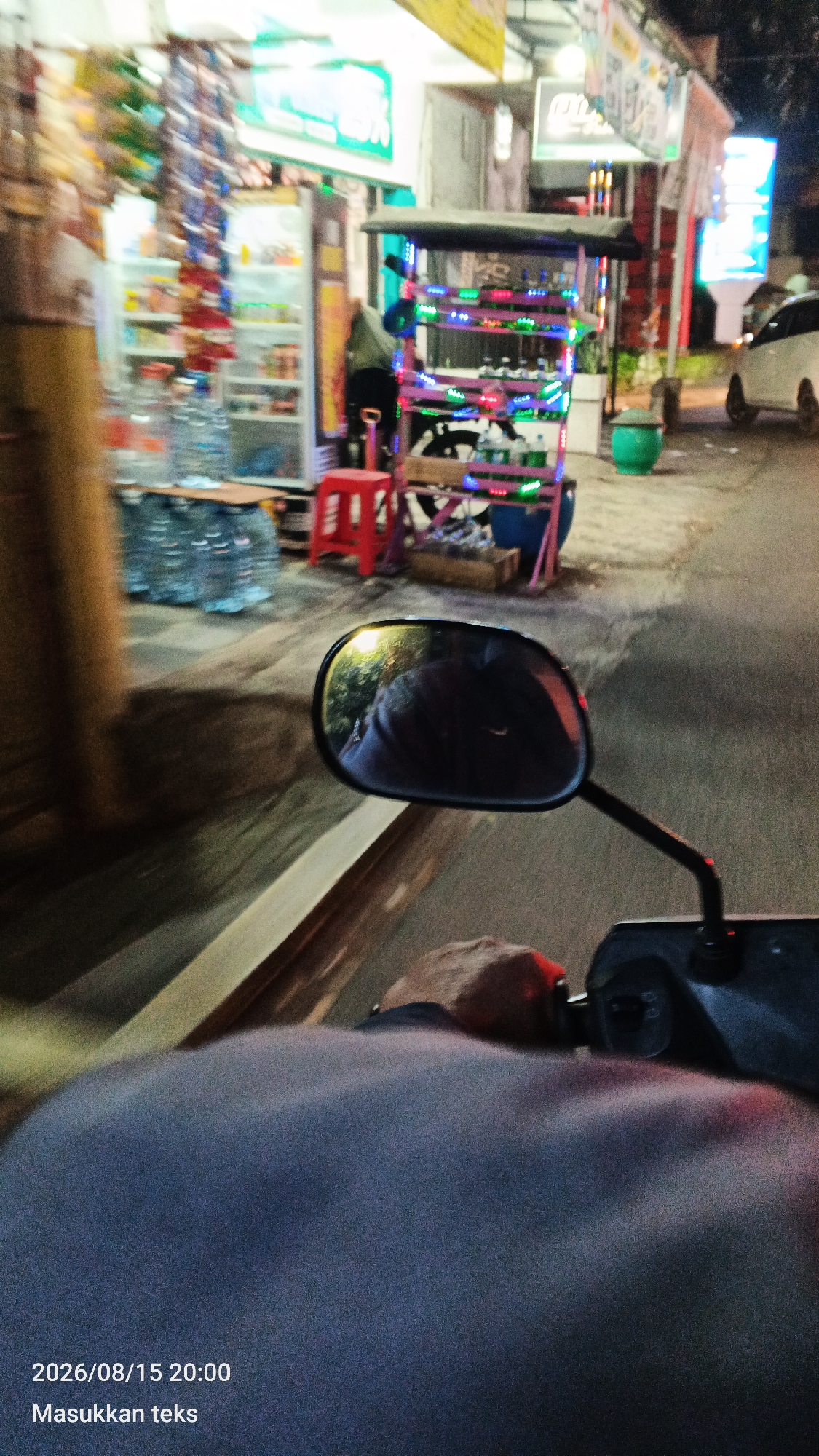 A first-person perspective shot from a motorcycle on a street at night in Malang, Indonesia. A person's head and shoulders are visible in the foreground, wearing a dark blue shirt. The motorcycle's side mirror is reflecting a person's face and trees. In the background, a small food cart decorated with colorful lights is visible, along with a red stool and several bottles of water. A refrigerator stocked with items and other stalls line the street. The street is paved, and there is a white car parked on the right side. The timestamp "2026/08/15 20:00" and the text "Masukkan teks" are overlaid on the bottom left of the image.