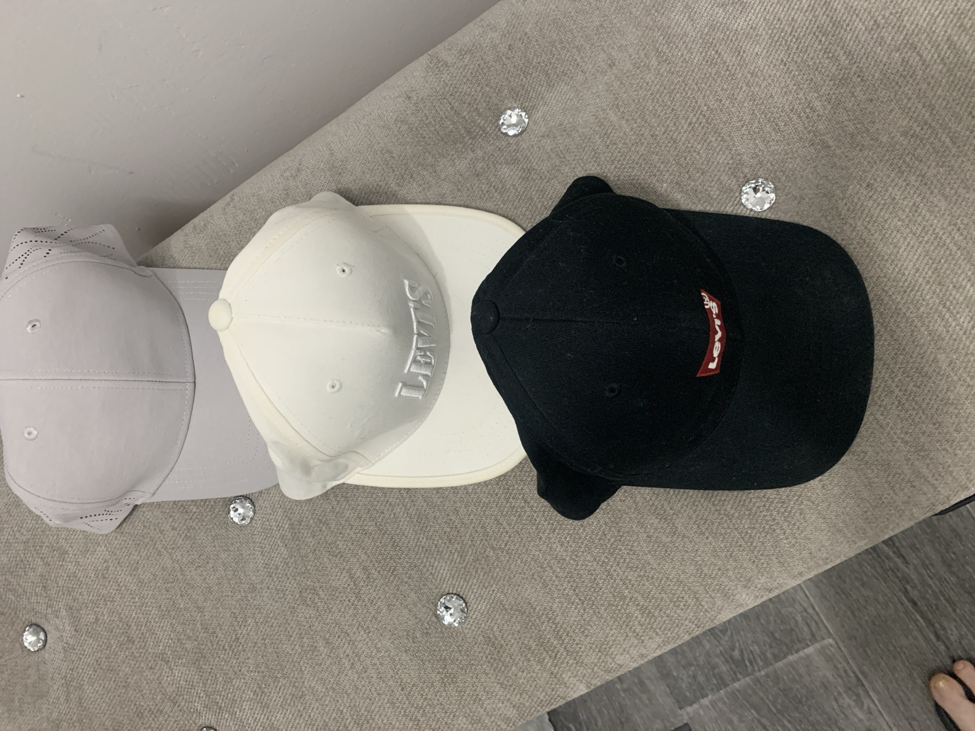 Three baseball caps are displayed on a textured, grey surface that appears to be a bench or ottoman, adorned with several clear, faceted embellishments resembling rhinestones. From left to right, the caps are light grey, white, and black. The light grey cap has a perforated crown. The white cap features raised, embossed lettering spelling "LEVIS". The black cap has a red "LEVIS" logo patch on its front. The overall scene suggests a casual display of accessories. The floor beneath the surface is visible on the right side of the image and appears to be made of grey wood-effect planks, suggesting an indoor setting. A foot with painted toenails is also visible at the bottom right corner, indicating the presence of a person. The lighting suggests it might be daytime, but no specific time of day can be definitively determined. The location context is Almaty, Kazakhstan, but there are no specific visual cues within the image to confirm this.