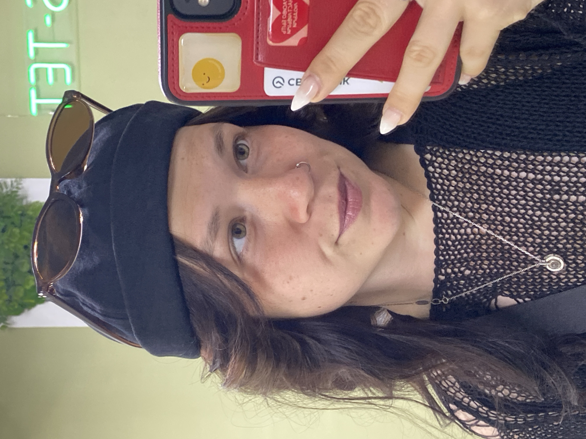 A young woman with long, dark, wavy hair and light brown eyes takes a selfie in a room. She is wearing a black beanie, sunglasses perched on top of the beanie, a black mesh top, and a silver necklace. She has a nose ring and her nails are painted with a French manicure. She is holding a red phone case in her right hand, which has a sticker of a smiling fried egg on it. In the background, a neon sign with Russian letters is visible, which appears to spell out "TET-2". The wall behind her is painted a light green color. The woman is positioned in the center of the frame, with her face slightly turned to the right. The overall impression is of a casual, indoor setting. The location is identified as Chaikovsky, Russia, based on the neon sign which likely refers to a specific establishment or location within the city.