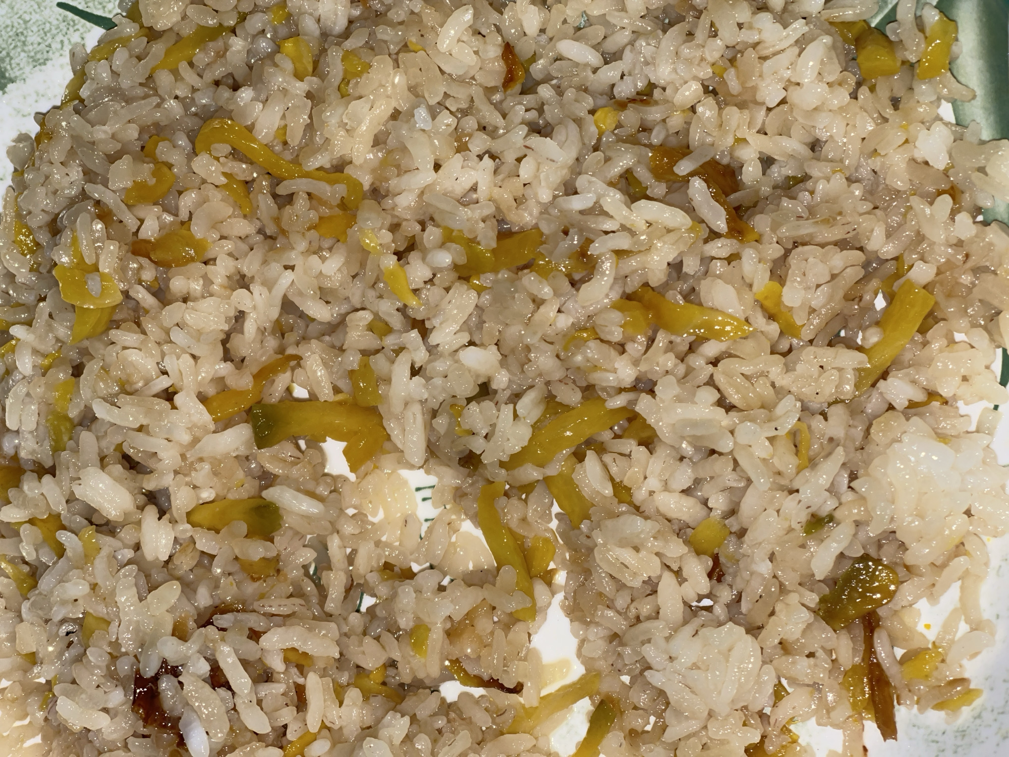 A close-up shot captures a plate of fluffy, white rice mixed with small, bright yellow pieces, possibly peppers or some other vegetable. The rice appears to be cooked and slightly oily, with individual grains distinct and glistening. The yellow pieces are cut into thin strips and are scattered throughout the rice, adding a vibrant contrast to the neutral color of the grains. The plate itself is partially visible at the edges of the frame, showing a decorative rim with a green and white patterned design. The lighting suggests an indoor setting, likely during the daytime, as there are no strong shadows. The overall impression is of a simple, prepared meal. No people or specific location cues are visible beyond the plate.