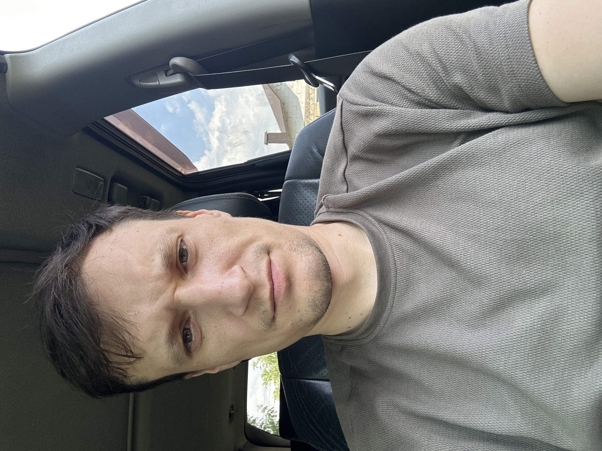 A man is sitting in the driver's seat of a car, looking directly at the camera. He has brown hair, light skin, and is wearing a taupe t-shirt. The interior of the car is visible, including a black leather seat and a section of the car's roof. Outside the car, through the window, a blue sky with white clouds and the roof of a building are visible. The image appears to be taken in Makiv, Ukraine, based on the building's architecture.