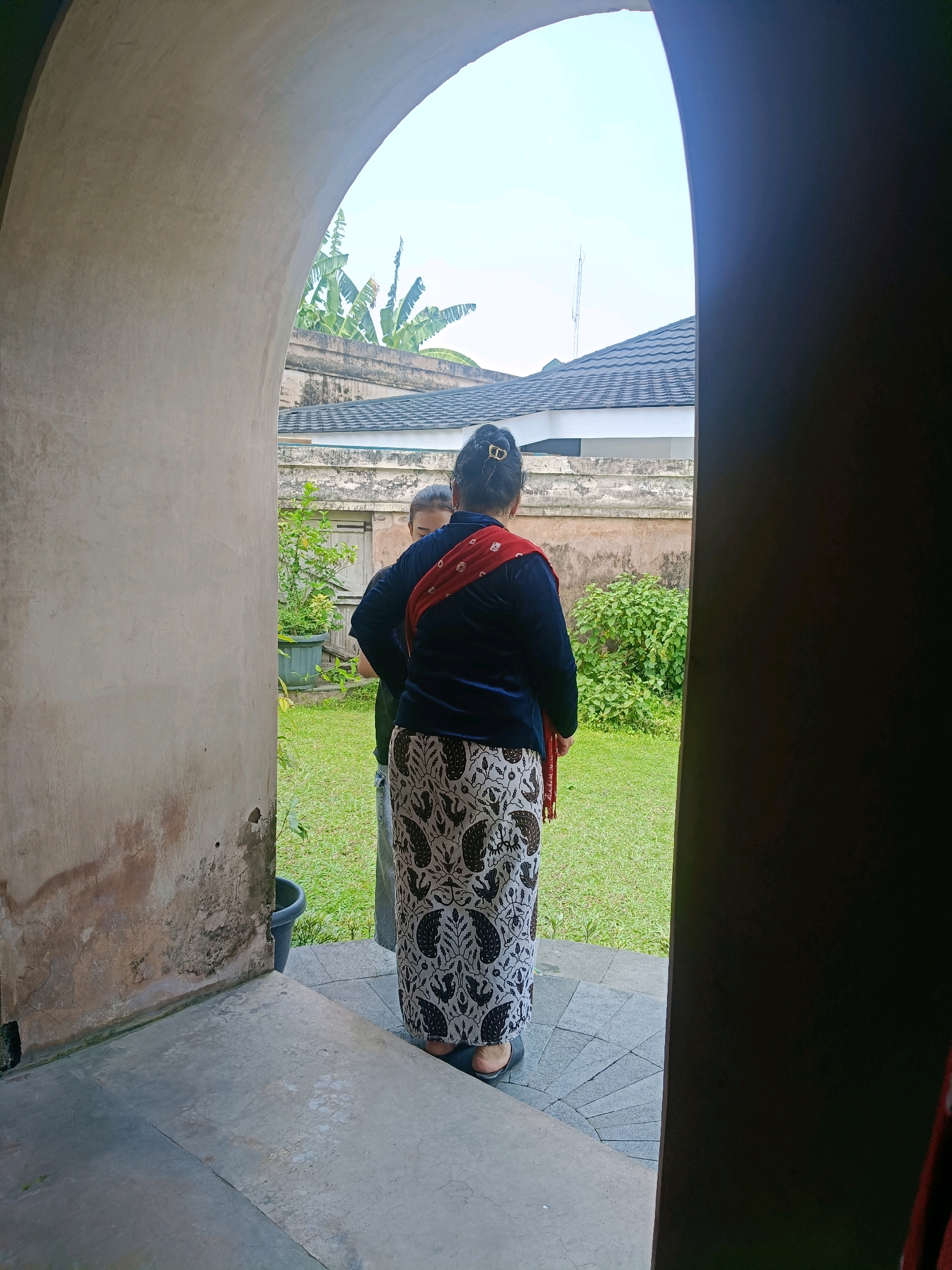 The image depicts a view through an arched entryway, looking out into a courtyard with lush green grass and several buildings in the background. In the foreground, two individuals are visible, facing away from the camera. The person closer to the foreground is wearing a dark blue jacket and a patterned sarong with white and black motifs. A red sash is draped over their shoulders. Their hair is pulled back into a bun, secured with a gold clip. The second individual, standing slightly behind and to the left, is wearing a green shirt and appears to be in conversation with the first person. The architecture suggests a historical or cultural site, possibly a palace or traditional dwelling, consistent with the architecture found in Yogyakarta, Indonesia. The sky is overcast, indicating a daytime scene. The entryway itself appears to be constructed of aged stone or concrete, showing signs of wear.