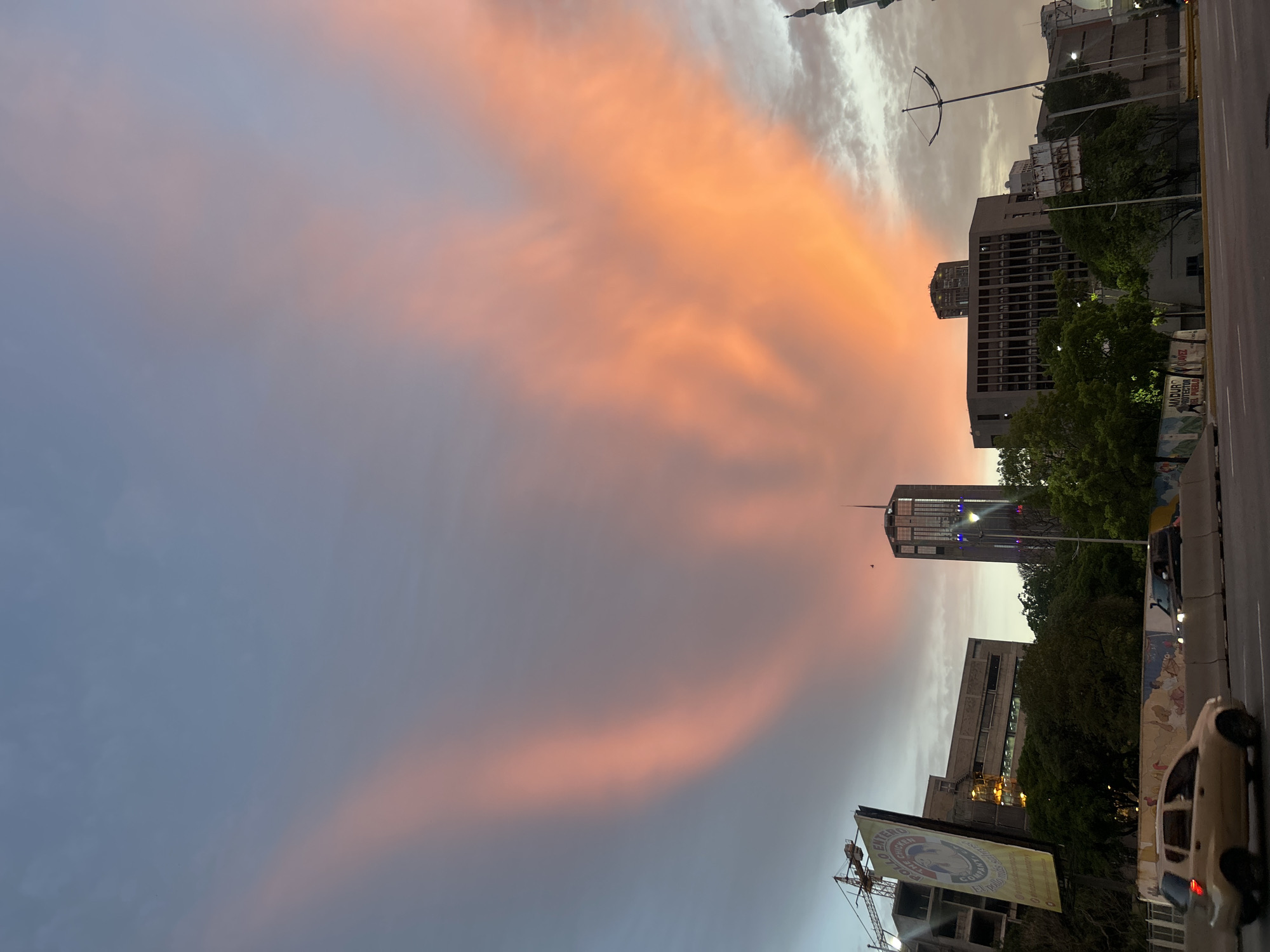 The image captures a sunset in Caracas, Venezuela, with prominent orange and pink clouds stretching across a gray-blue sky. Several buildings of varying heights are visible, including a tall skyscraper with lit-up blue and white lights. A street runs along the right side of the image, with a cream-colored car in the foreground. A billboard, with a crane visible behind it, stands out in the lower right corner. Green trees are scattered throughout the scene, interspersed with the buildings. The lighting indicates that it is dusk.