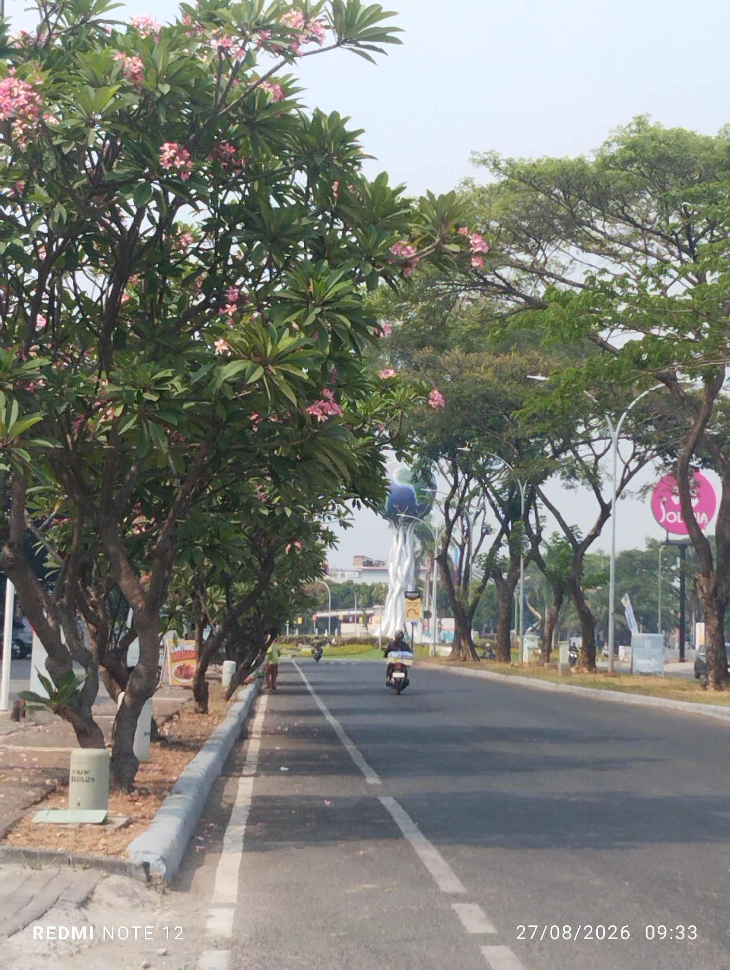 The image depicts a street scene in East Jakarta, Indonesia, on a clear day. A row of trees with pink flowers lines the left side of a paved road. Some of the trees are young, with thin trunks, while others are mature and have thicker branches. The road is empty of cars but has a few motorcycles visible. One motorcycle is parked on the side of the road near a concrete barrier, and another is riding away from the camera in the distance. There are buildings visible in the background, including a large, white, abstract sculpture. The sky is overcast, and the lighting suggests it is late morning or early afternoon. The image is watermarked with "REDMI NOTE 12" in the bottom left corner and the date and time "27/08/2026 09:33" in the bottom right corner.