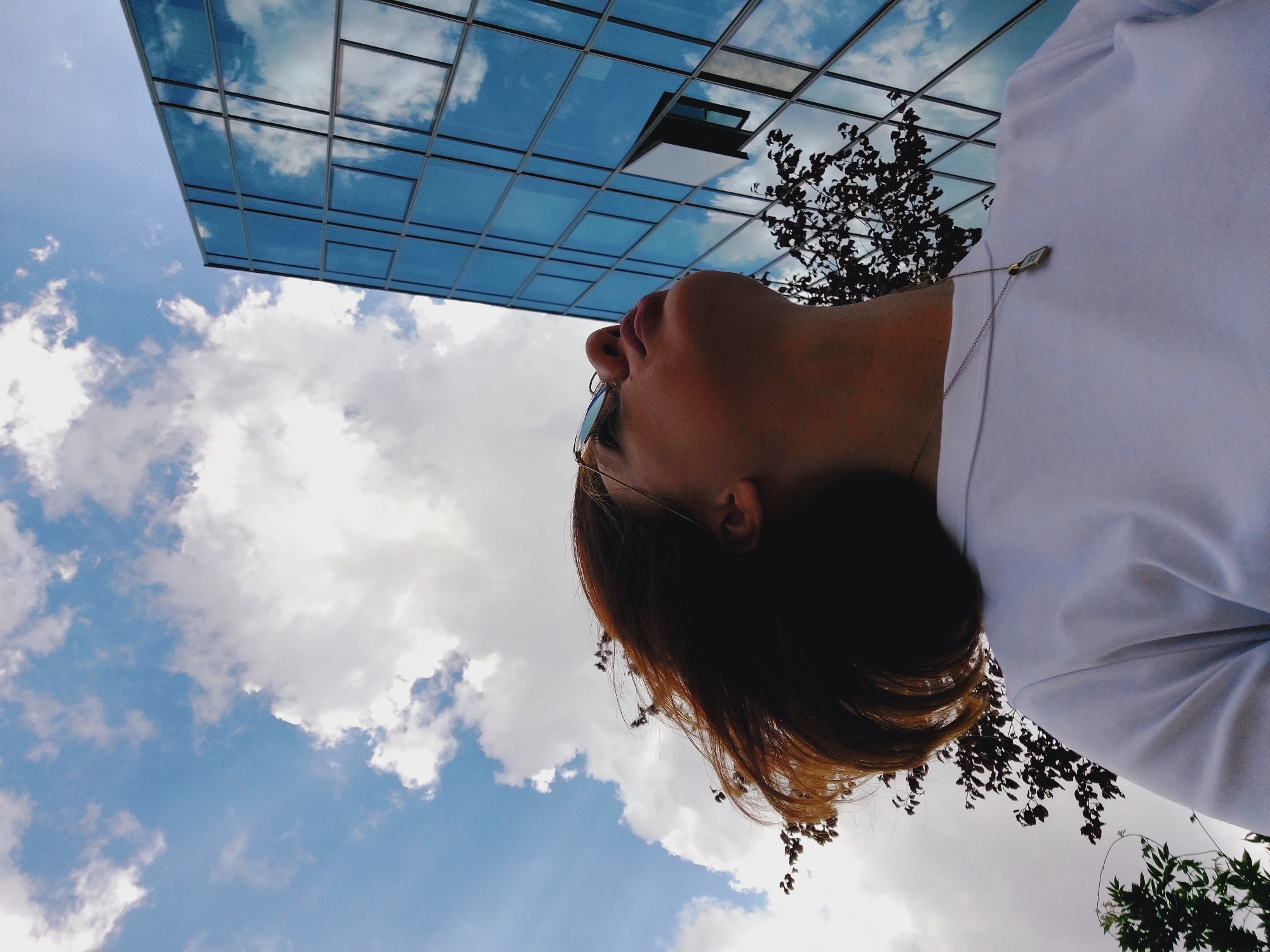 In Sofia, Bulgaria, a young woman with short, reddish-brown hair, wearing sunglasses and a white t-shirt, looks up towards the sky. She is positioned in the foreground, with her head tilted back and her profile visible. Behind her, a modern building with a glass facade dominates the upper right portion of the frame, its reflective surface mirroring the sky and clouds. Trees with dark foliage are visible in the middle ground, interspersed between the woman and the building. The sky above is a mix of blue with scattered white clouds, some of which are illuminated by sunlight, creating a slightly dramatic effect. The overall impression is one of a moment of contemplation or observation in an urban environment.