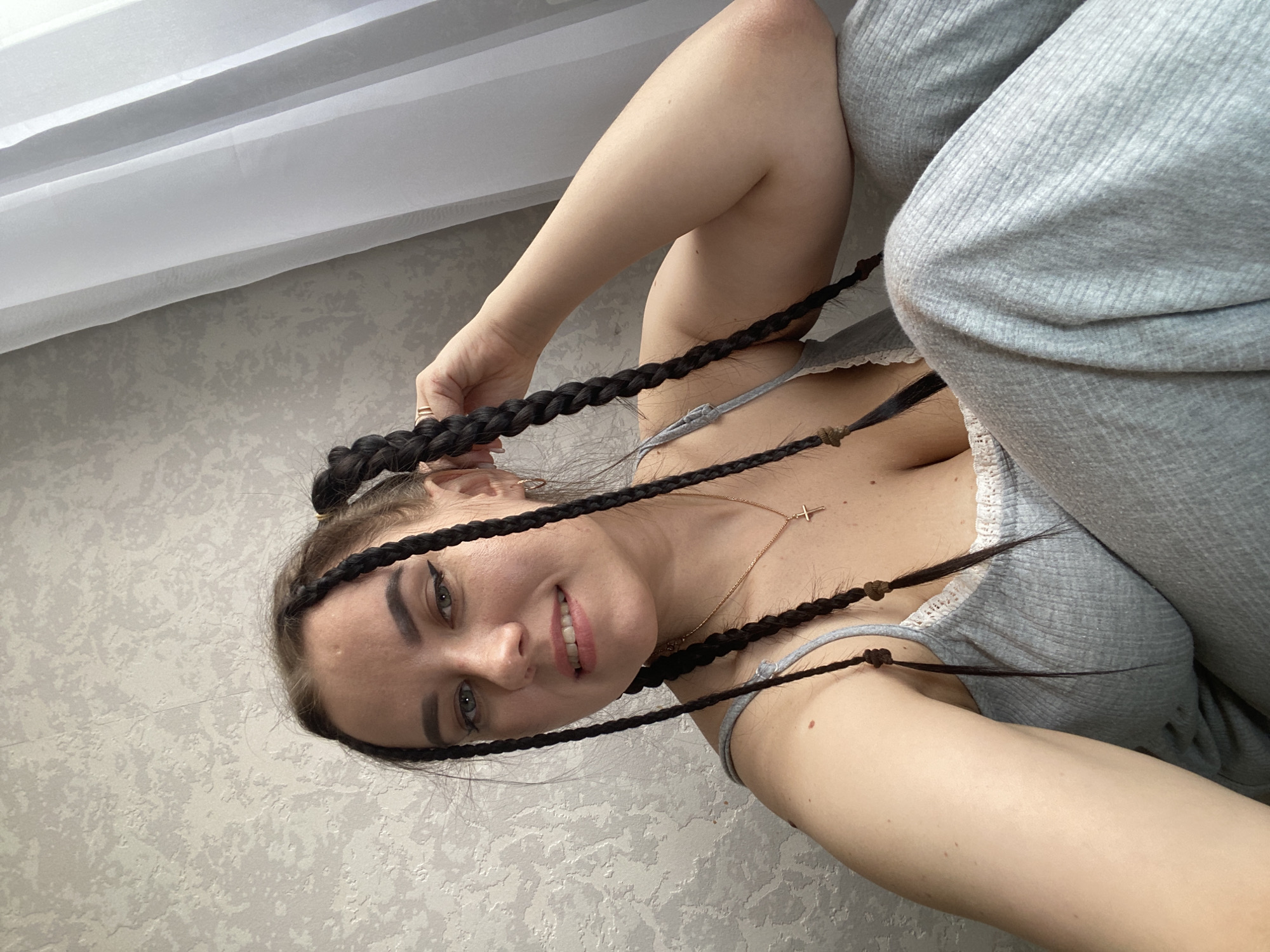 A fair-skinned woman with braided hair and dark eyeliner smiles at the camera. She wears a light gray top and pants. Her left hand is partially visible, holding one of her braids. A gold necklace with a cross pendant is visible on her chest. She appears to be indoors, with a white curtain and textured gray wallpaper as the background. There is no specific information provided indicating this image was taken in the city of Chaikovsky, Russia.