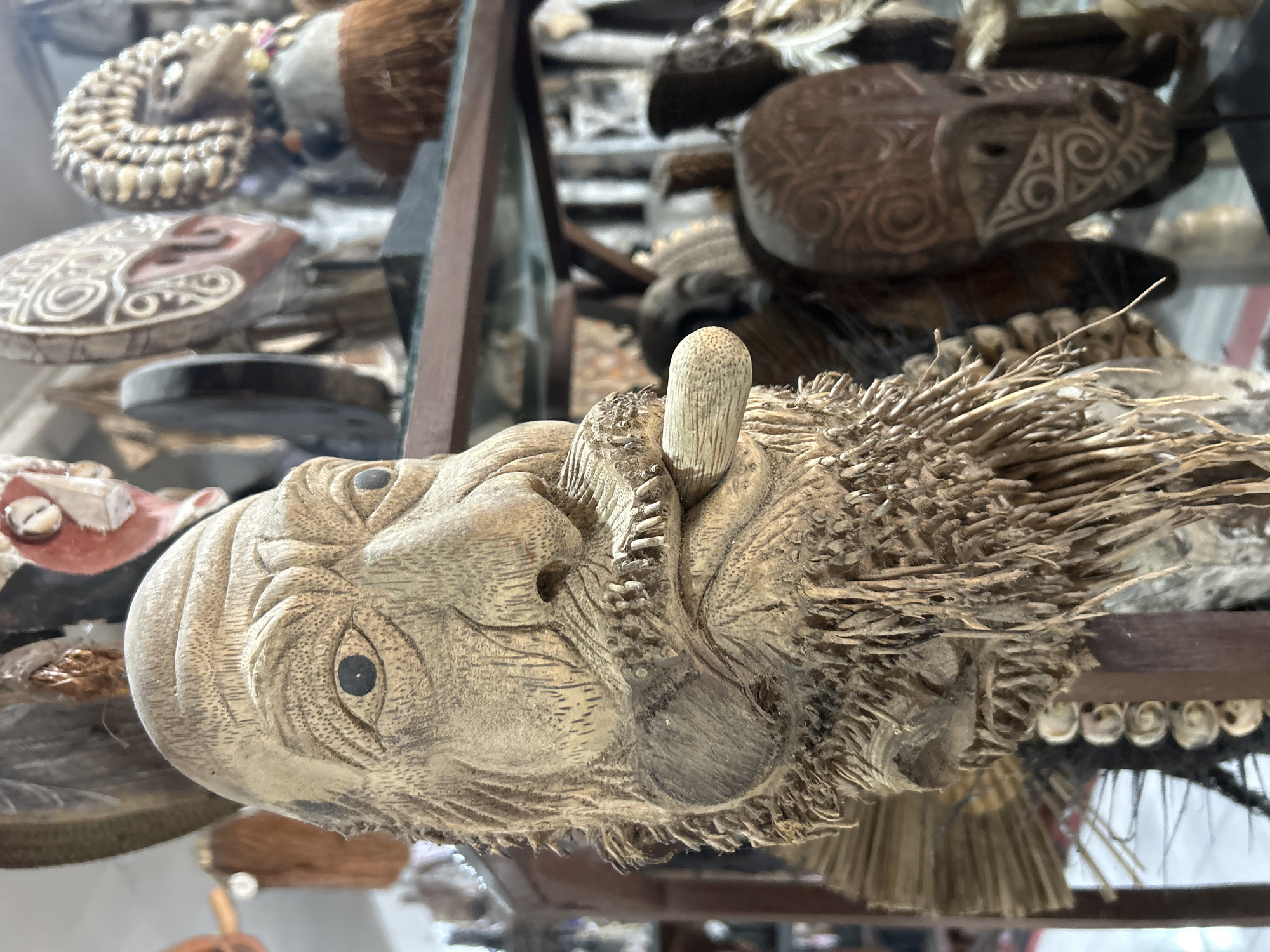 A close-up, eye-level, outdoor shot focuses on a carved wooden mask that resembles an elderly man with a beard and a cigar or pipe in his mouth. The mask is positioned in the foreground, slightly off-center to the left, and is the primary subject. Its textured surface shows intricate details of wrinkles, a prominent nose, and a beard made of dried plant material or strings. The eyes are dark and appear to be made of a different material, possibly obsidian or a dark stone.
In the background, out of focus, are other decorative masks and objects displayed on shelves, suggesting a market or shop setting. These include several tribal masks with distinct patterns and materials, such as shells and feathers. The lighting is soft and diffused, casting no harsh shadows. The overall impression is that of a cultural artifact display in a marketplace. The specific location is given as Unknown, Unknown.